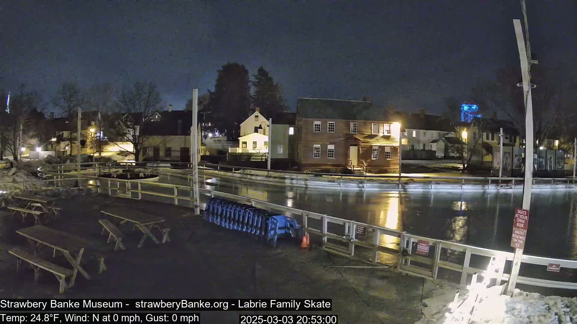 An outdoor ice rink at night under a clear sky, surrounded by buildings and picnic tables, with calm conditions.