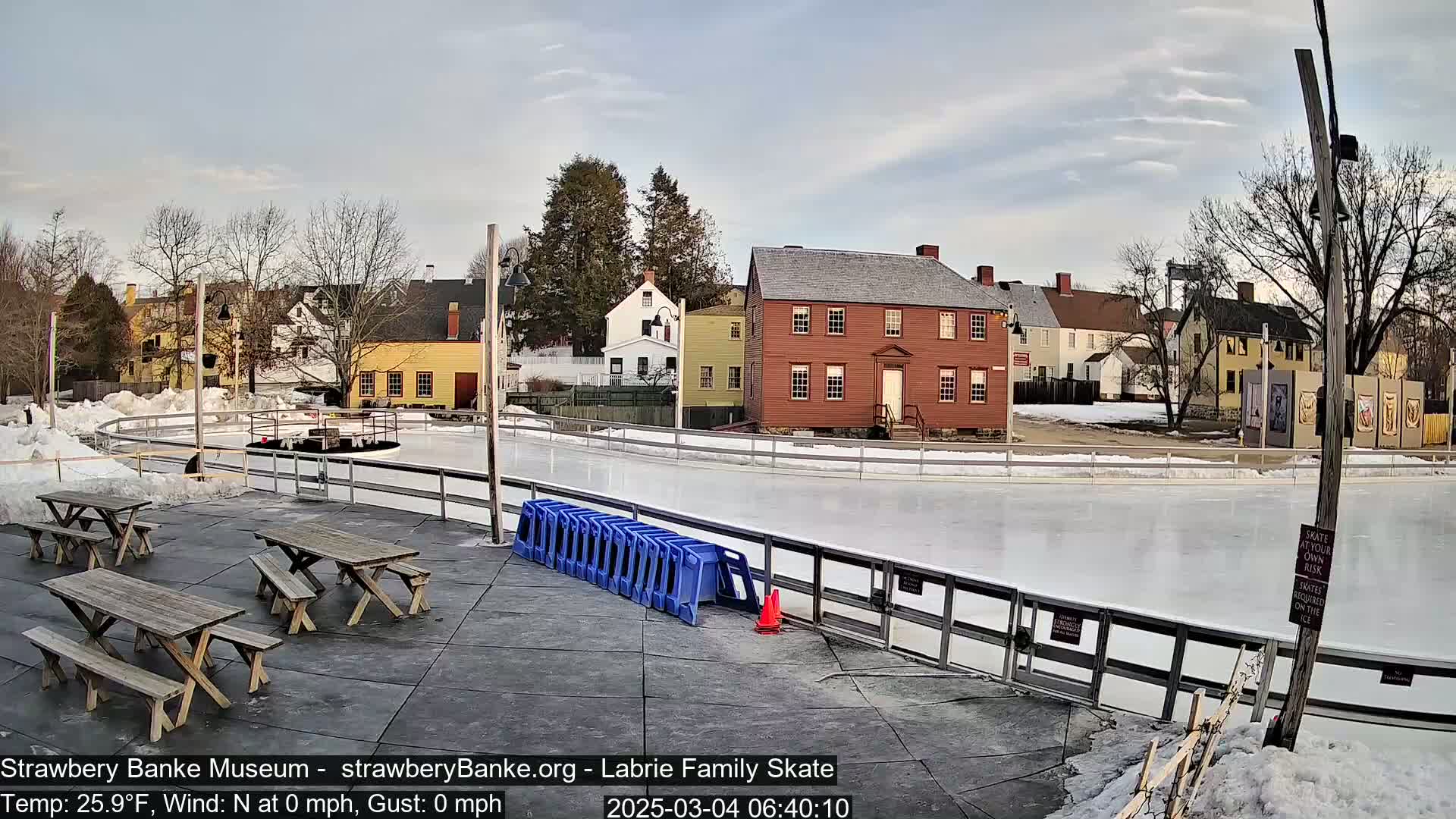 An outdoor ice skating rink, surrounded by historic buildings and picnic tables, on a partly cloudy day.