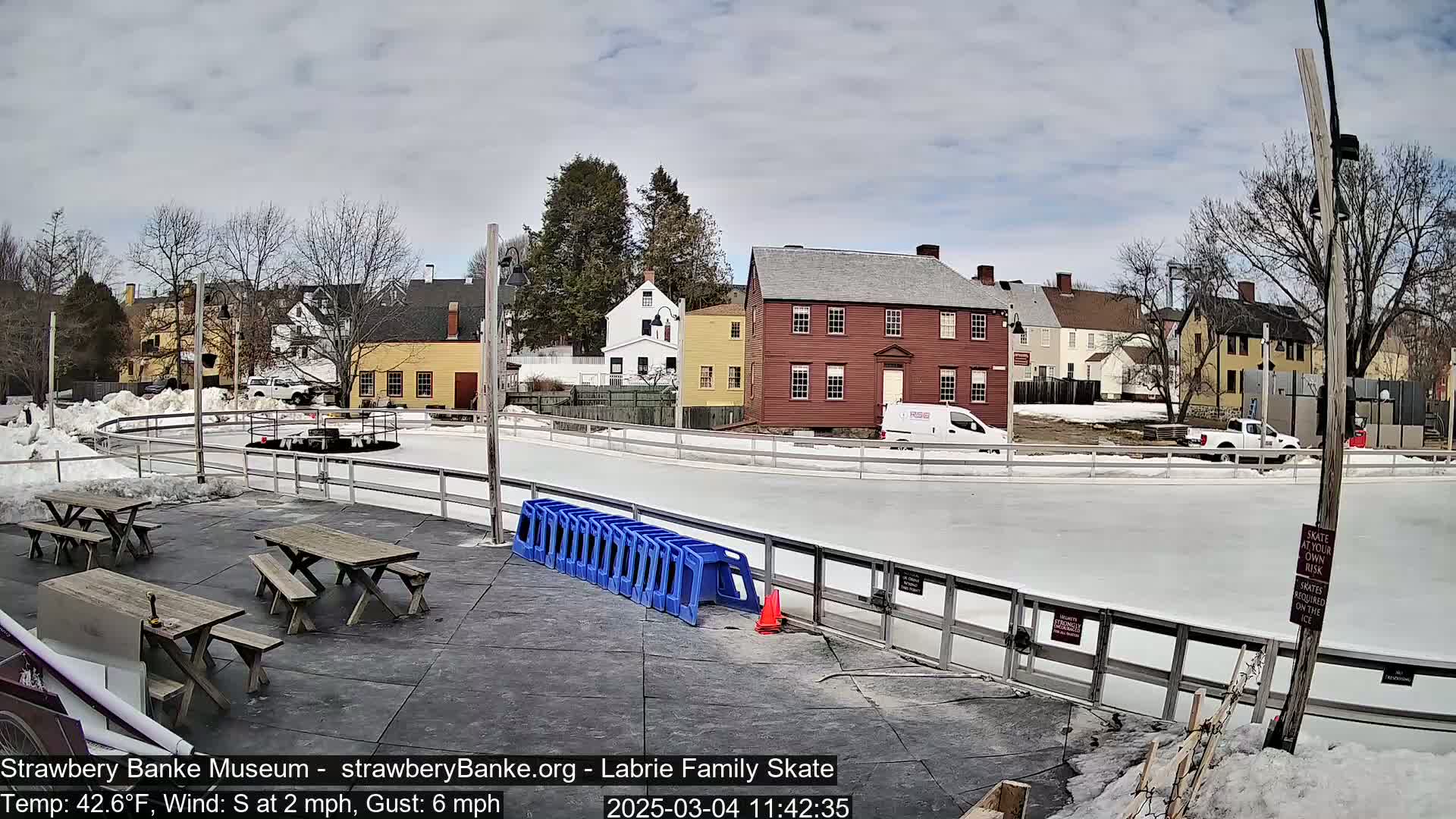 An outdoor ice skating rink, surrounded by snow and historic buildings, on a partly cloudy day with a south wind.