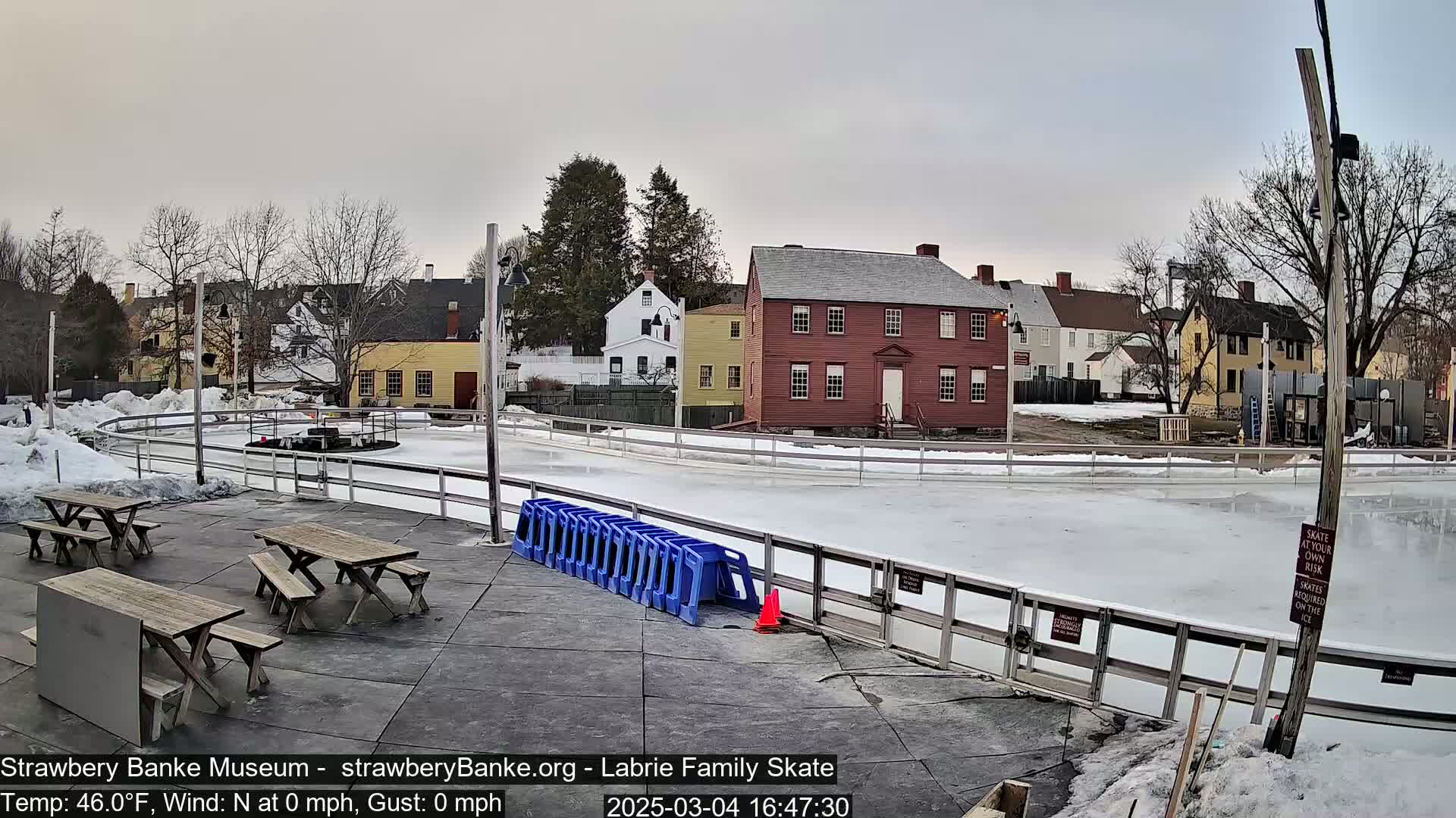 An outdoor ice skating rink is surrounded by historical-looking buildings under a cloudy sky, with several picnic tables and blue plastic barriers nearby.