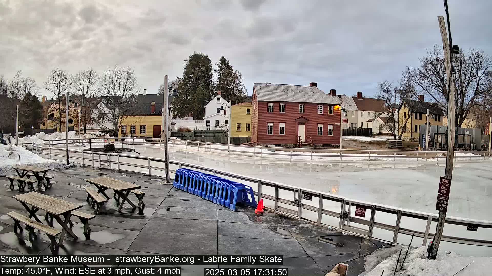 An outdoor ice rink surrounded by historic buildings on an overcast day, with several picnic tables nearby.
