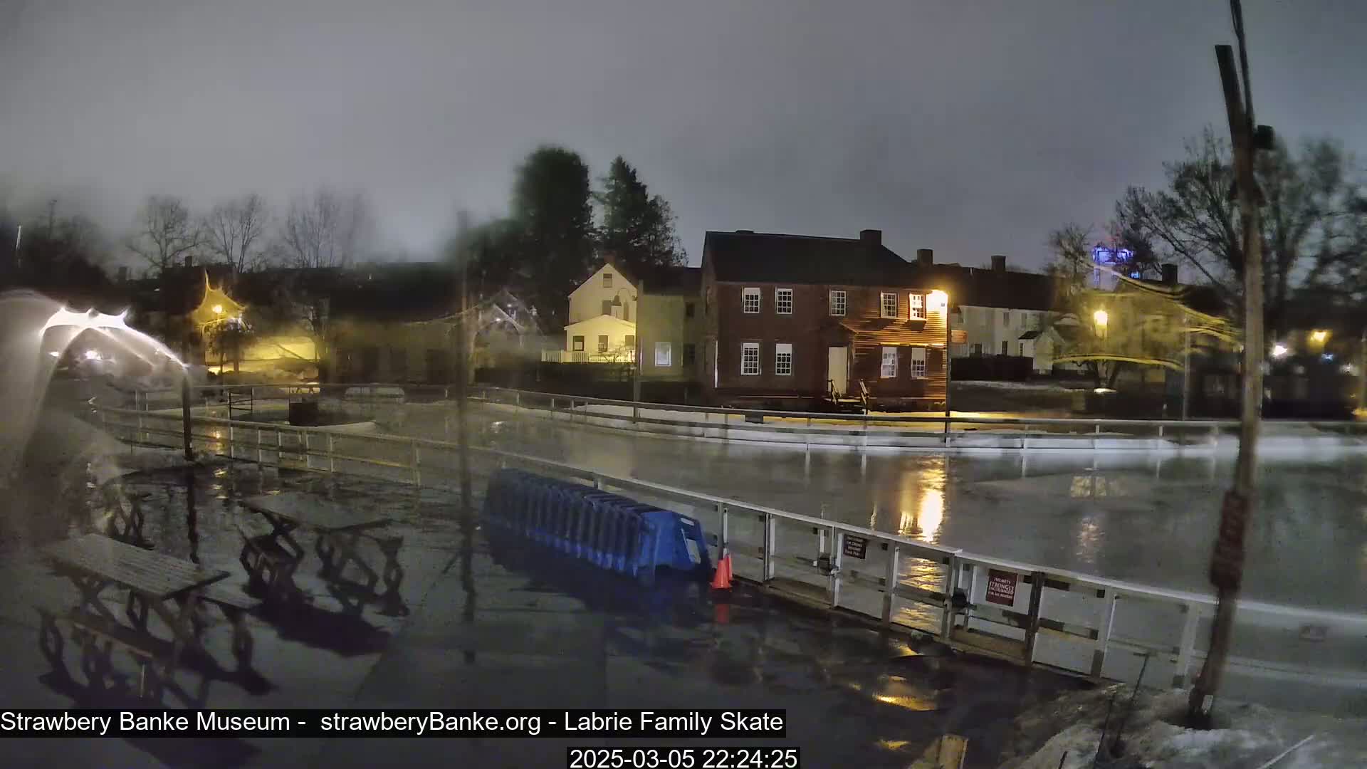 A rain-soaked outdoor ice rink sits in front of a row of historic buildings under a dark, overcast night sky.