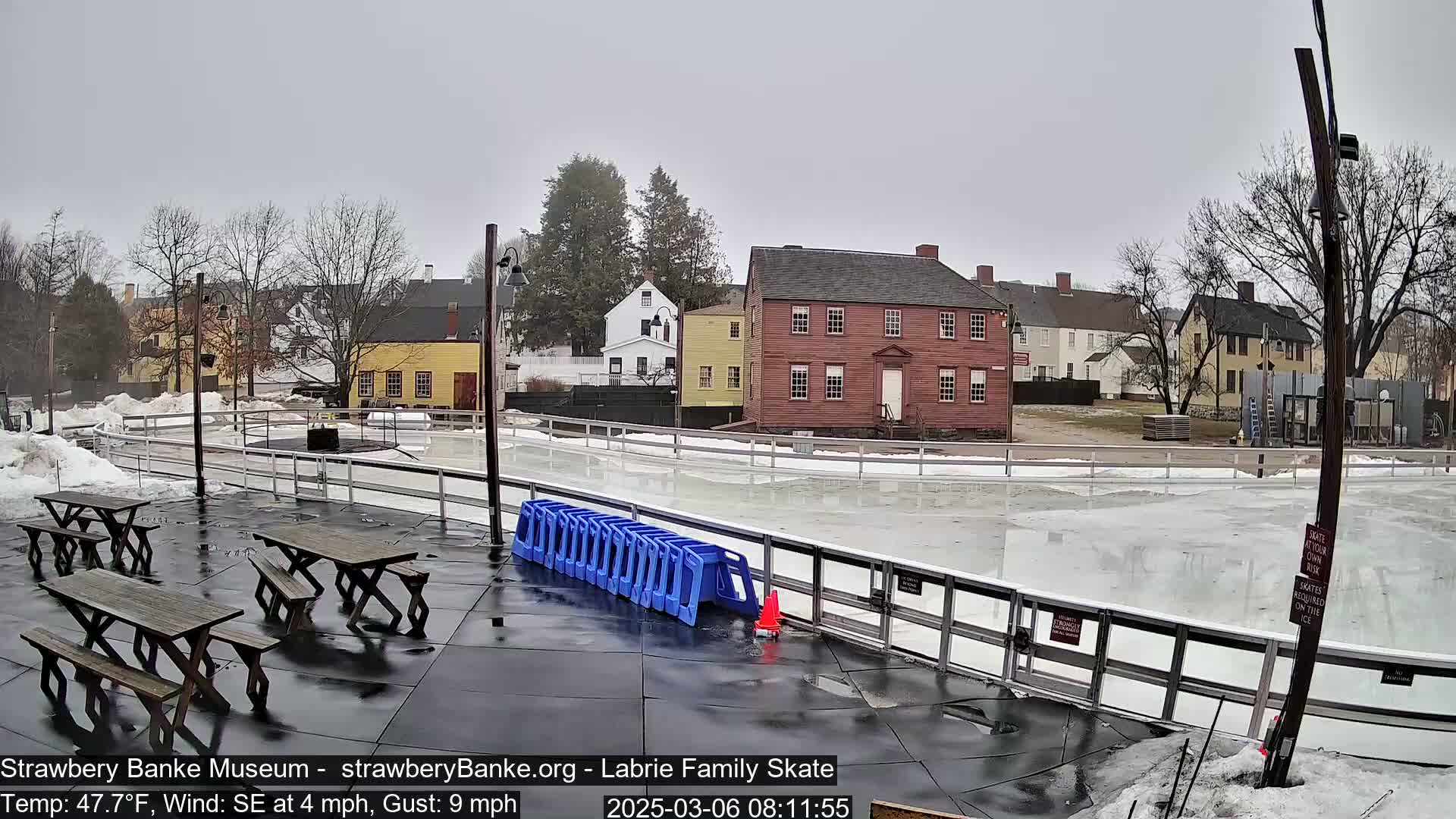 A mostly empty outdoor ice rink is surrounded by several colonial-style buildings under a gray, overcast sky.