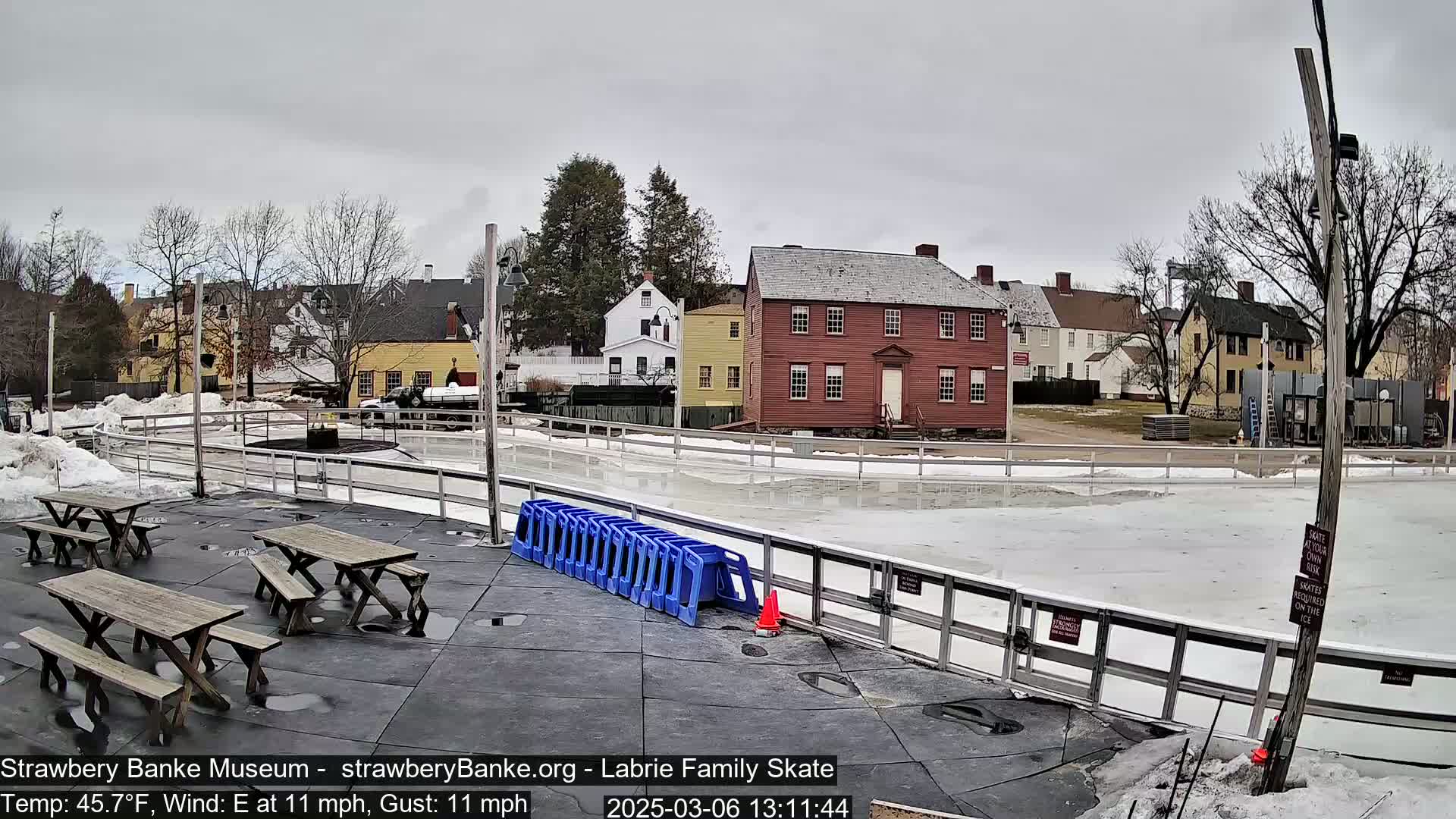 An outdoor ice skating rink with picnic tables and a row of blue plastic chairs sits in front of several historic-looking buildings on an overcast day with an east wind.