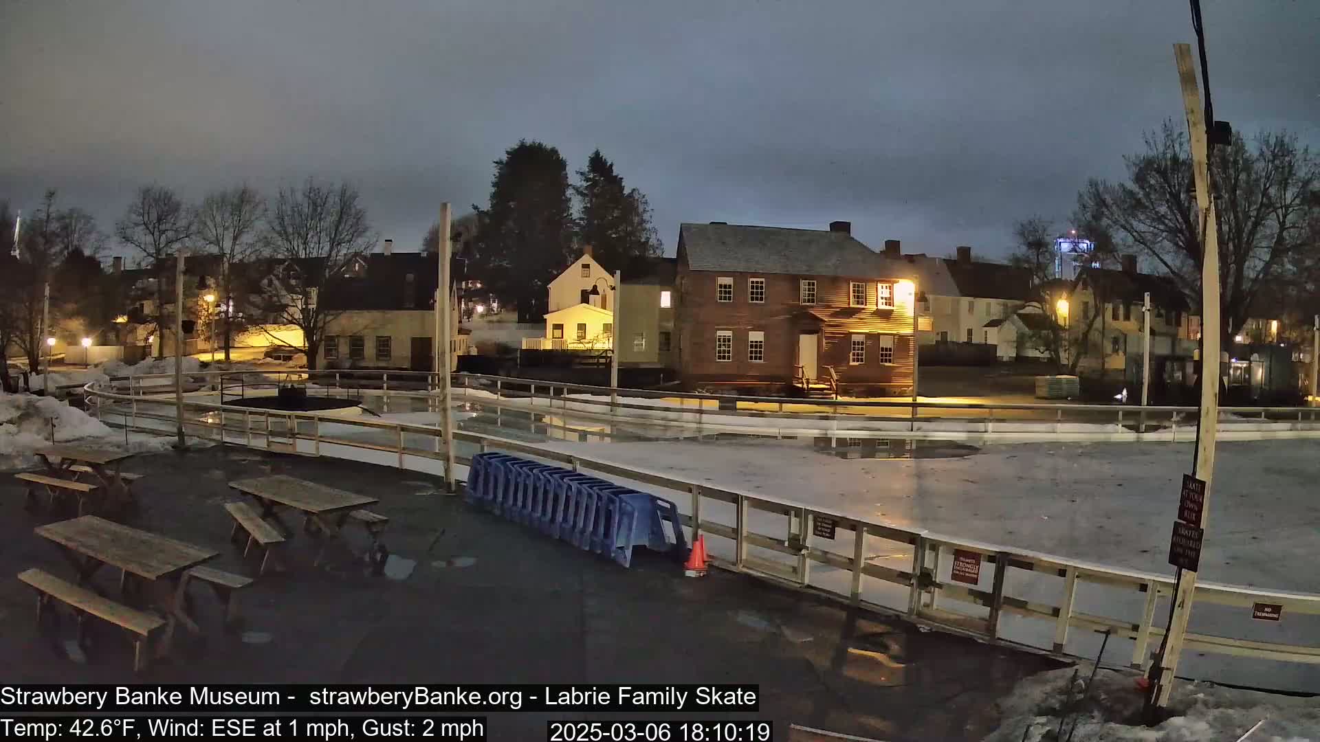An outdoor ice skating rink at night, partially covered in ice and surrounded by historic buildings under a dark, cloudy sky.