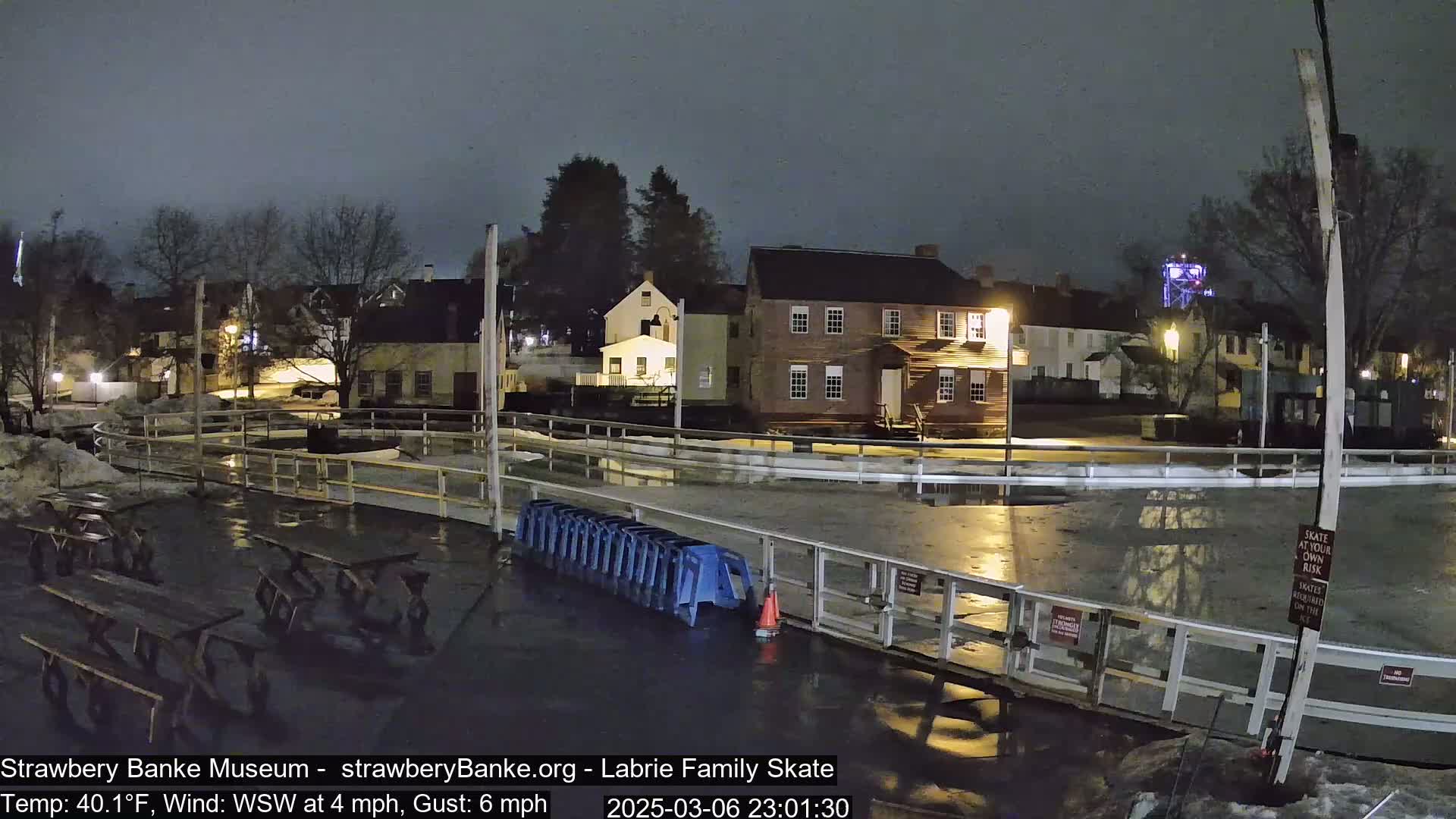 A nighttime view of an outdoor ice rink, partially surrounded by historic-looking buildings under a dark sky, with a light wind.