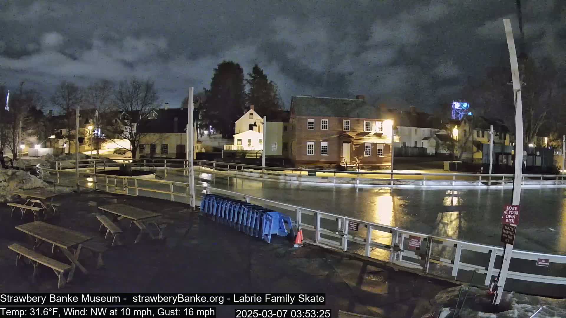 At night, under a cloudy sky, a wet outdoor ice rink is surrounded by historical-looking buildings and picnic tables.