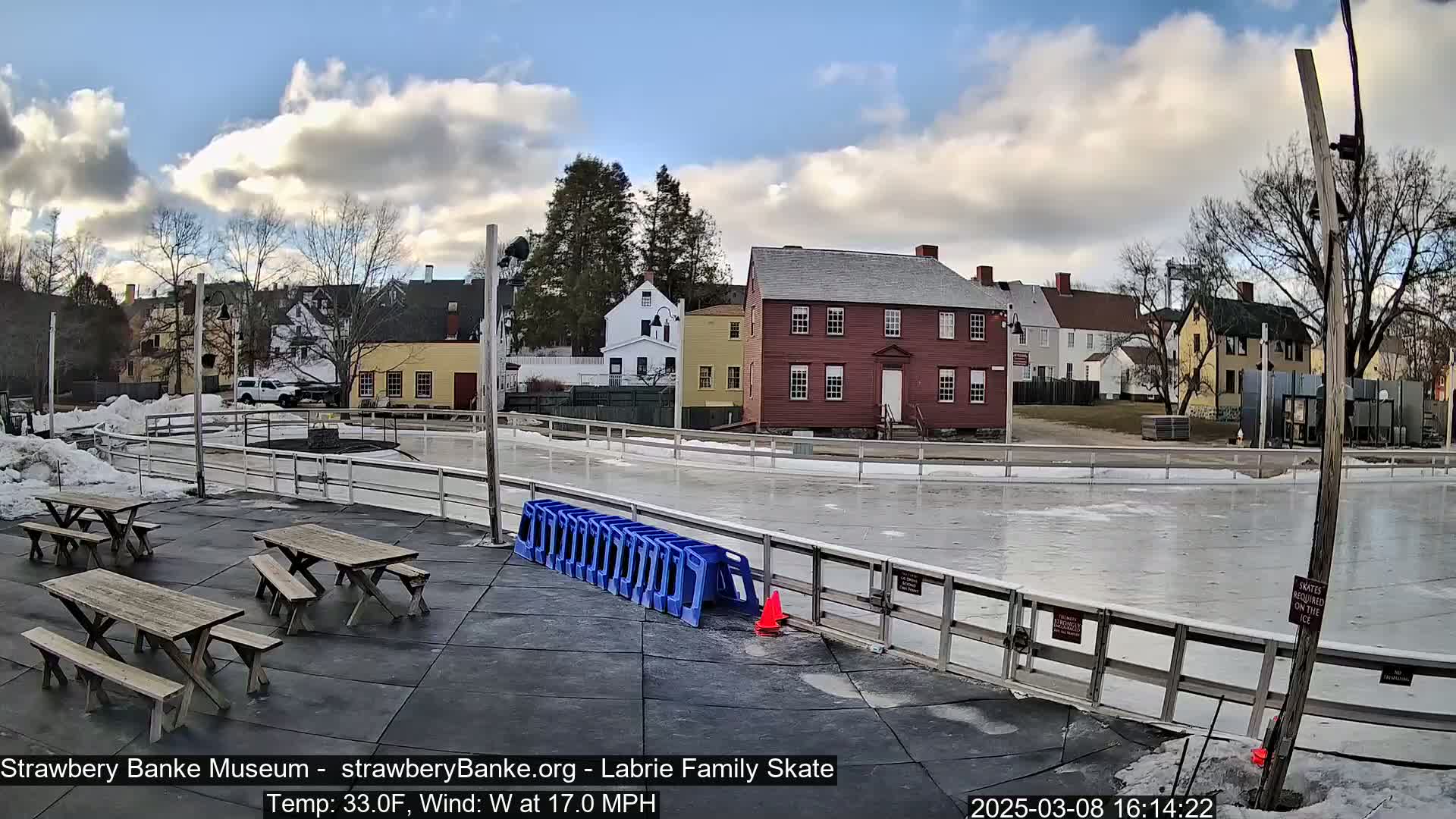An outdoor ice skating rink with picnic tables sits in front of several historic-looking buildings under a partly cloudy sky.