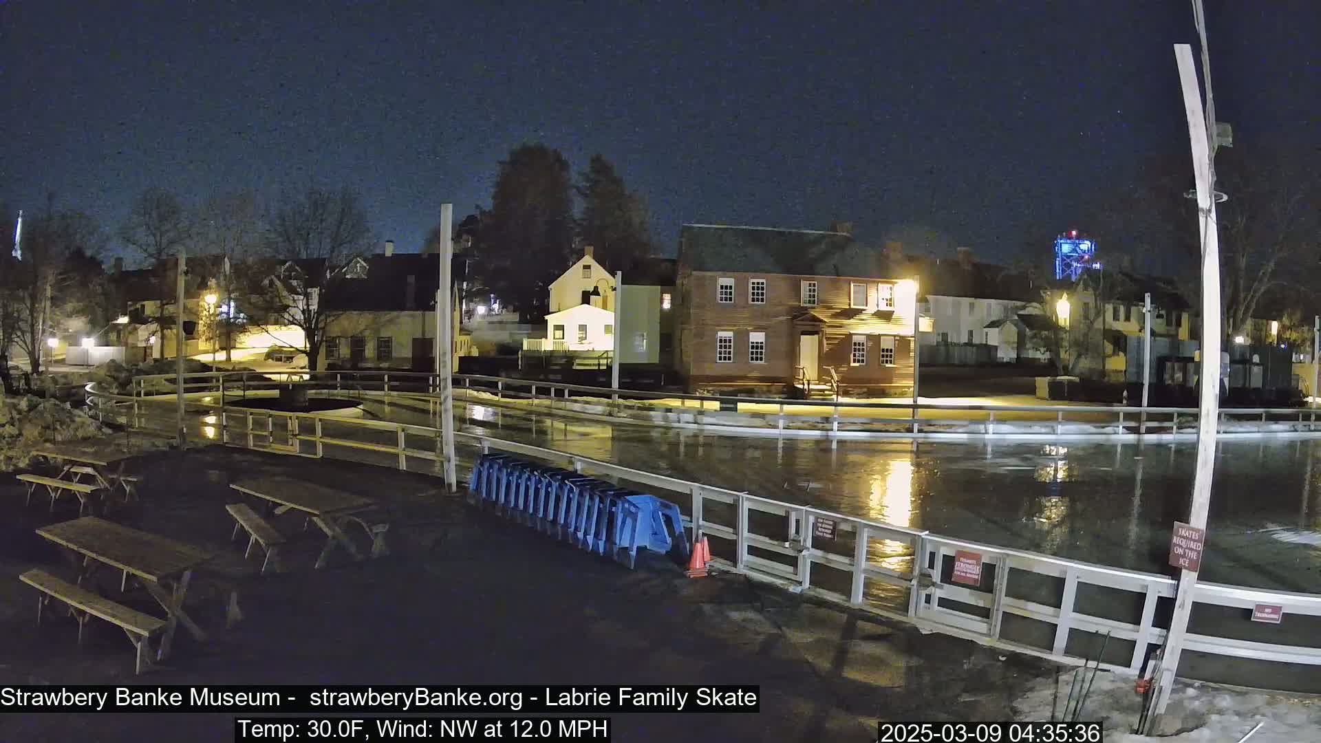A nighttime view of a flooded outdoor skating rink surrounded by historic buildings under a clear, dark sky, with a northwest wind of 12 mph and a temperature of 30 degrees Fahrenheit.
