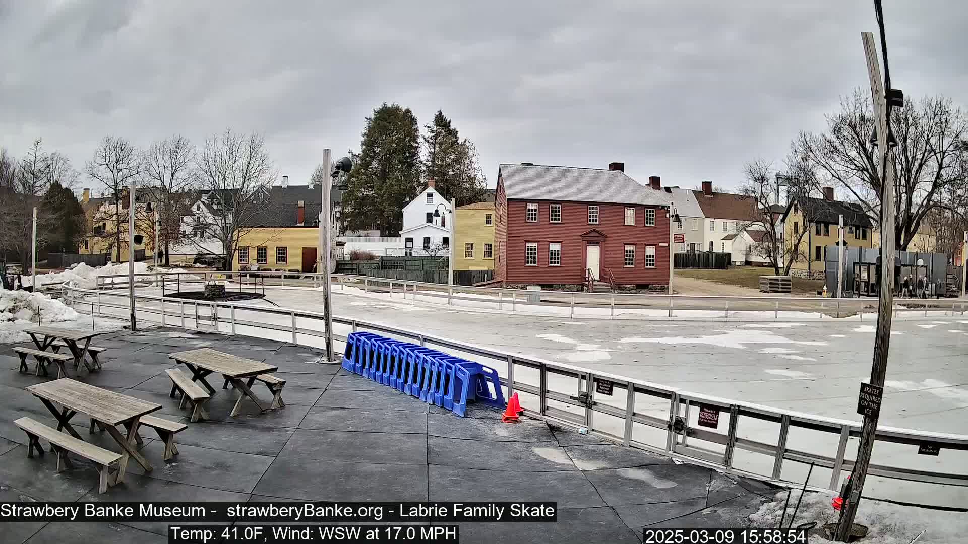 An outdoor ice rink with picnic tables is situated in front of several historic-looking buildings under a cloudy sky.