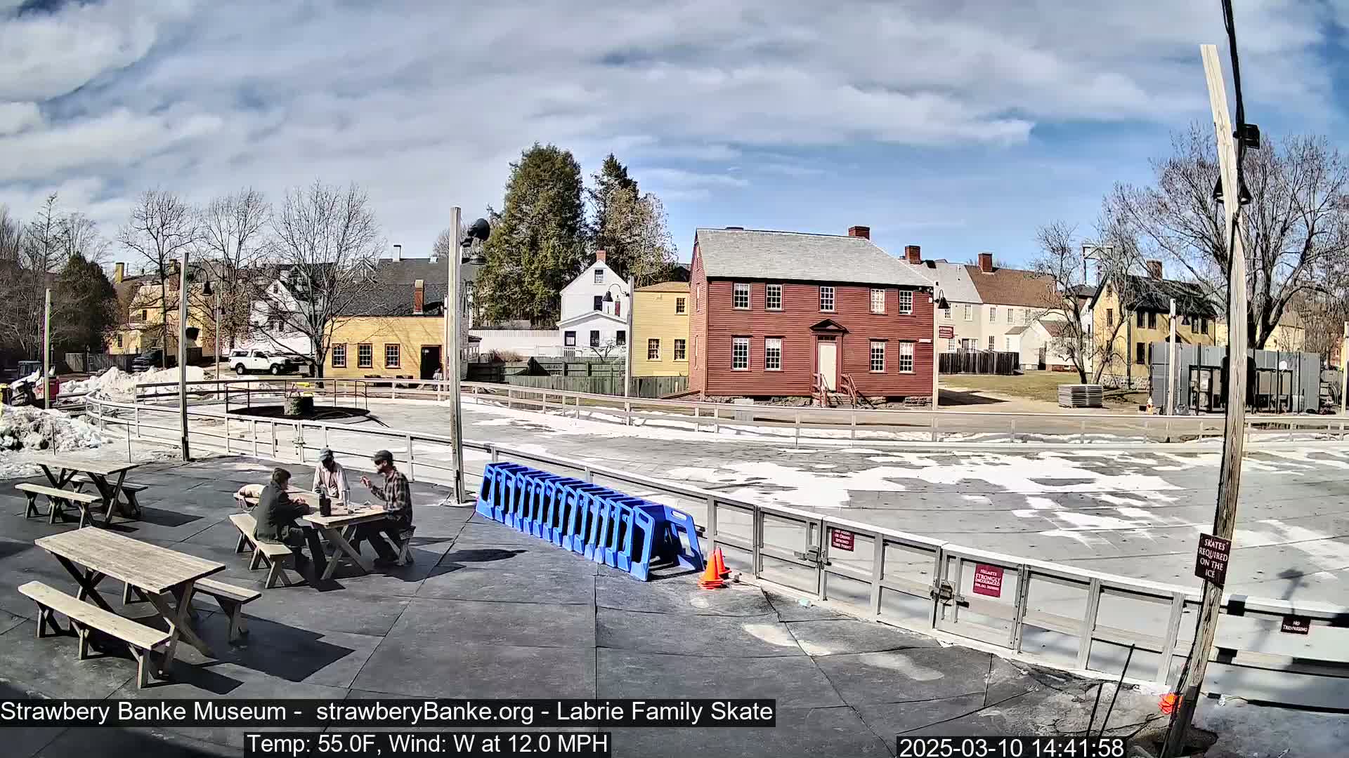 Three people sit at a picnic table near an outdoor skating rink surrounded by historical buildings on a partly sunny day with a westerly wind.