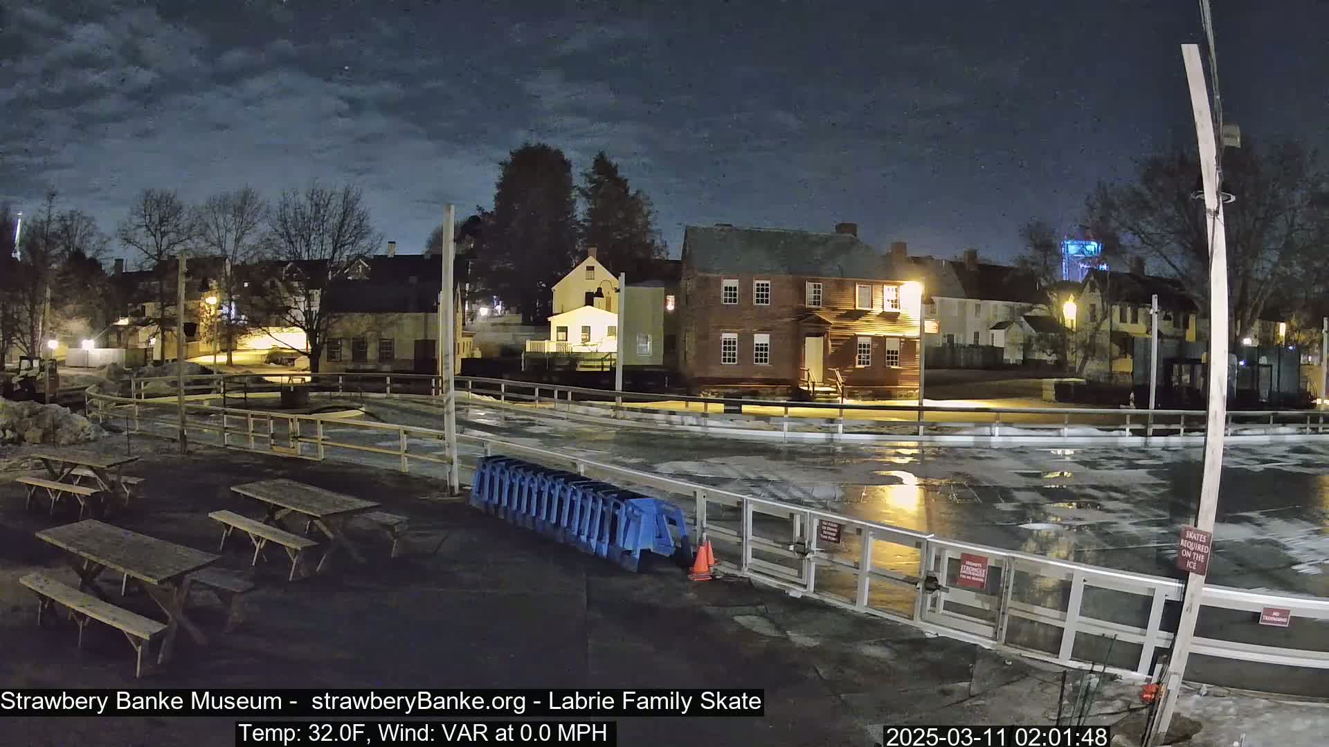 An outdoor nighttime scene shows a partially-melted ice skating rink surrounded by several picnic tables, a wooden fence, and historic-looking buildings under a partly cloudy sky.