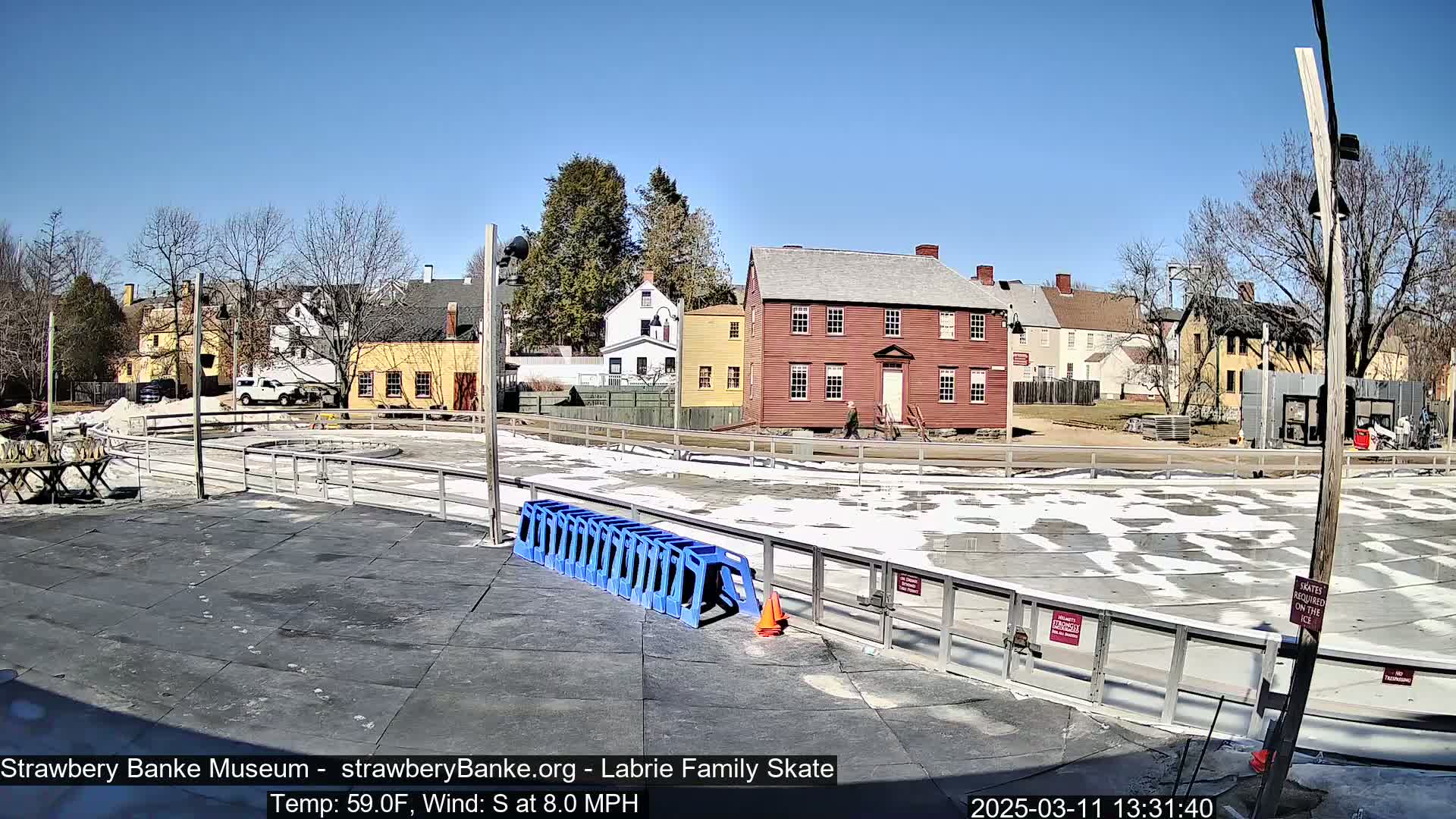 A mostly melted outdoor ice rink is situated in front of several historic-looking buildings on a sunny day.