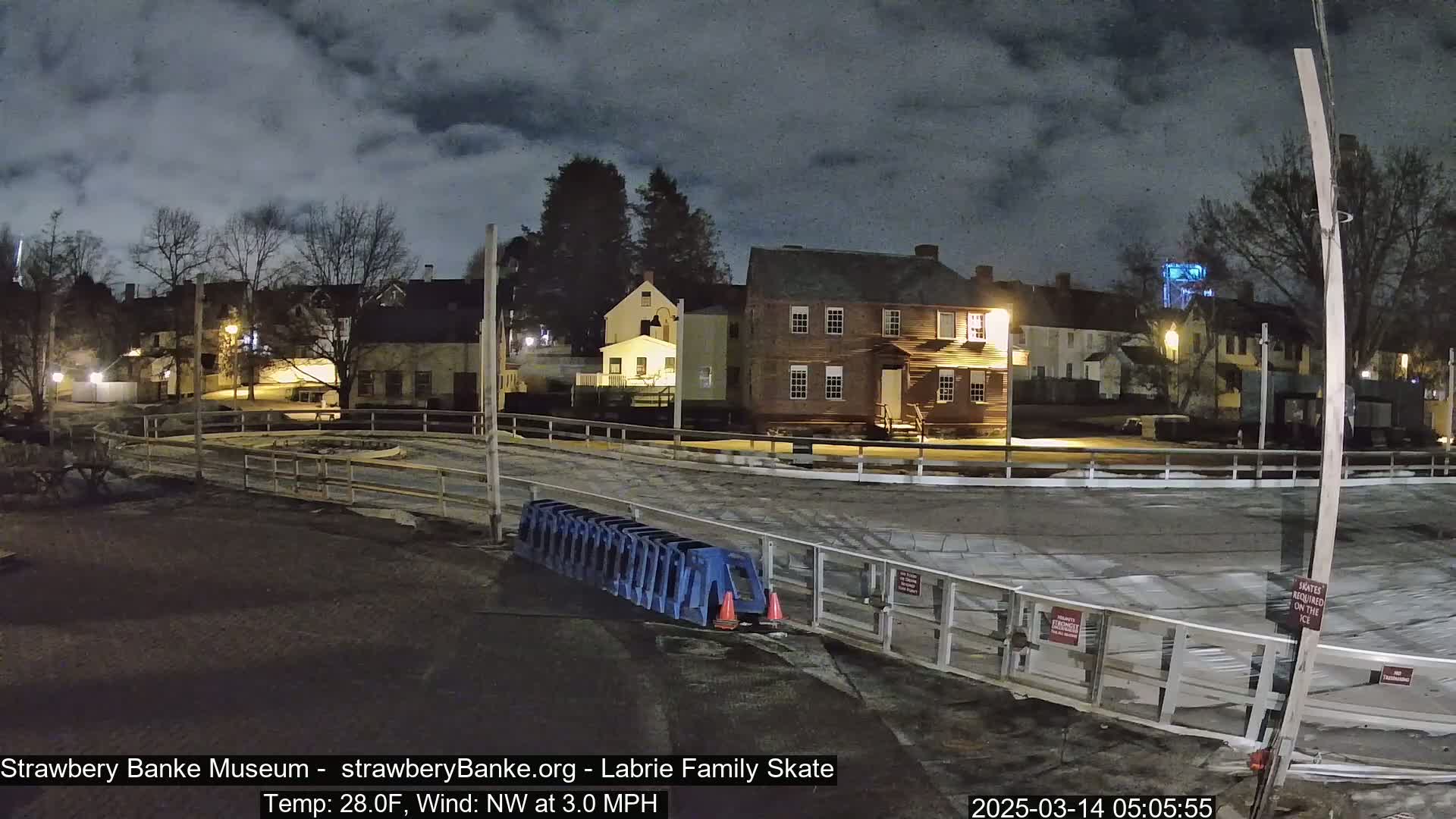 A nighttime view of an outdoor ice rink surrounded by historic-looking buildings under a cloudy sky.