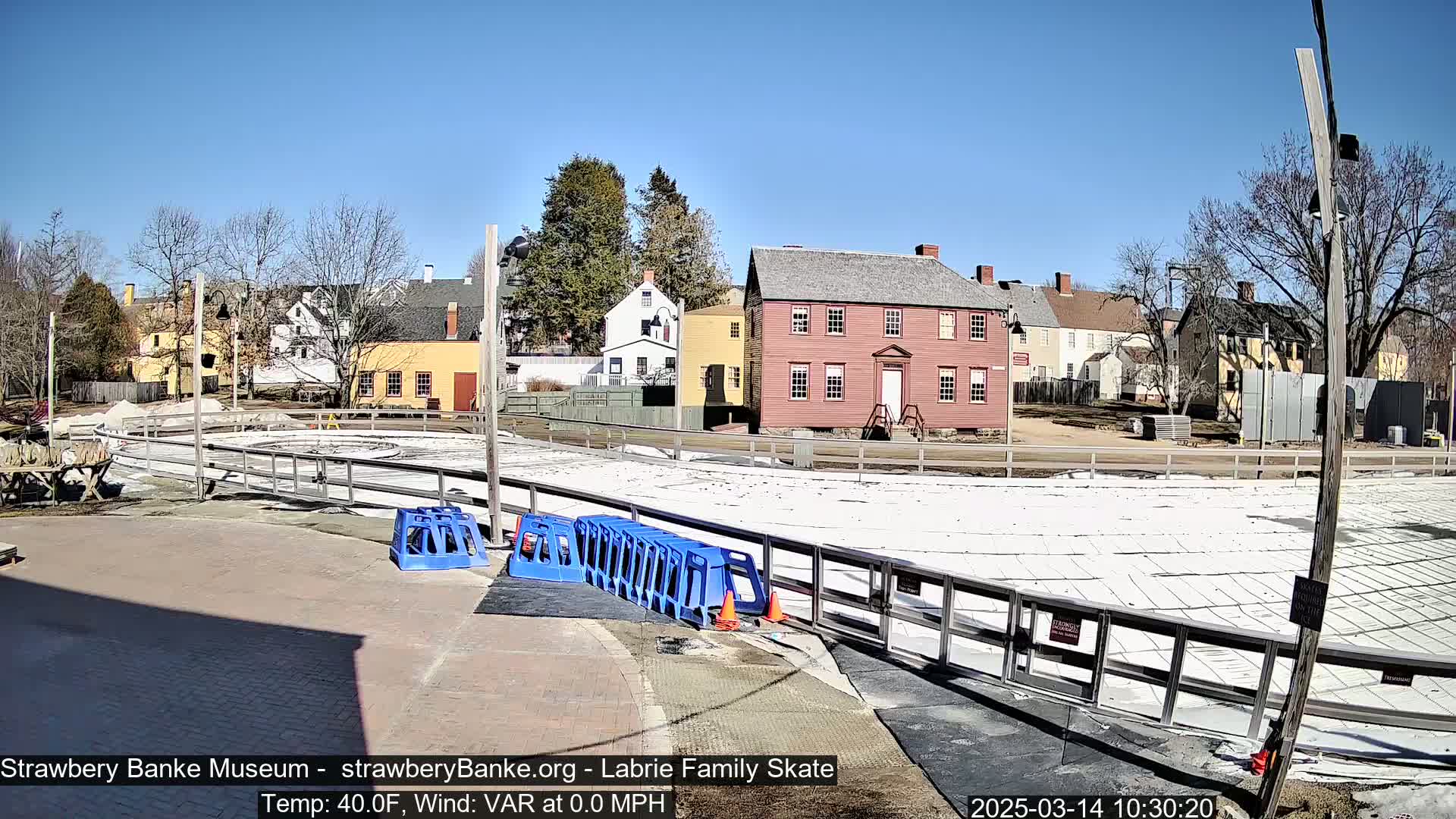 A mostly clear, sunny day reveals an outdoor skating rink surrounded by historical-looking buildings, with blue plastic barriers and orange cones near the rink's edge.