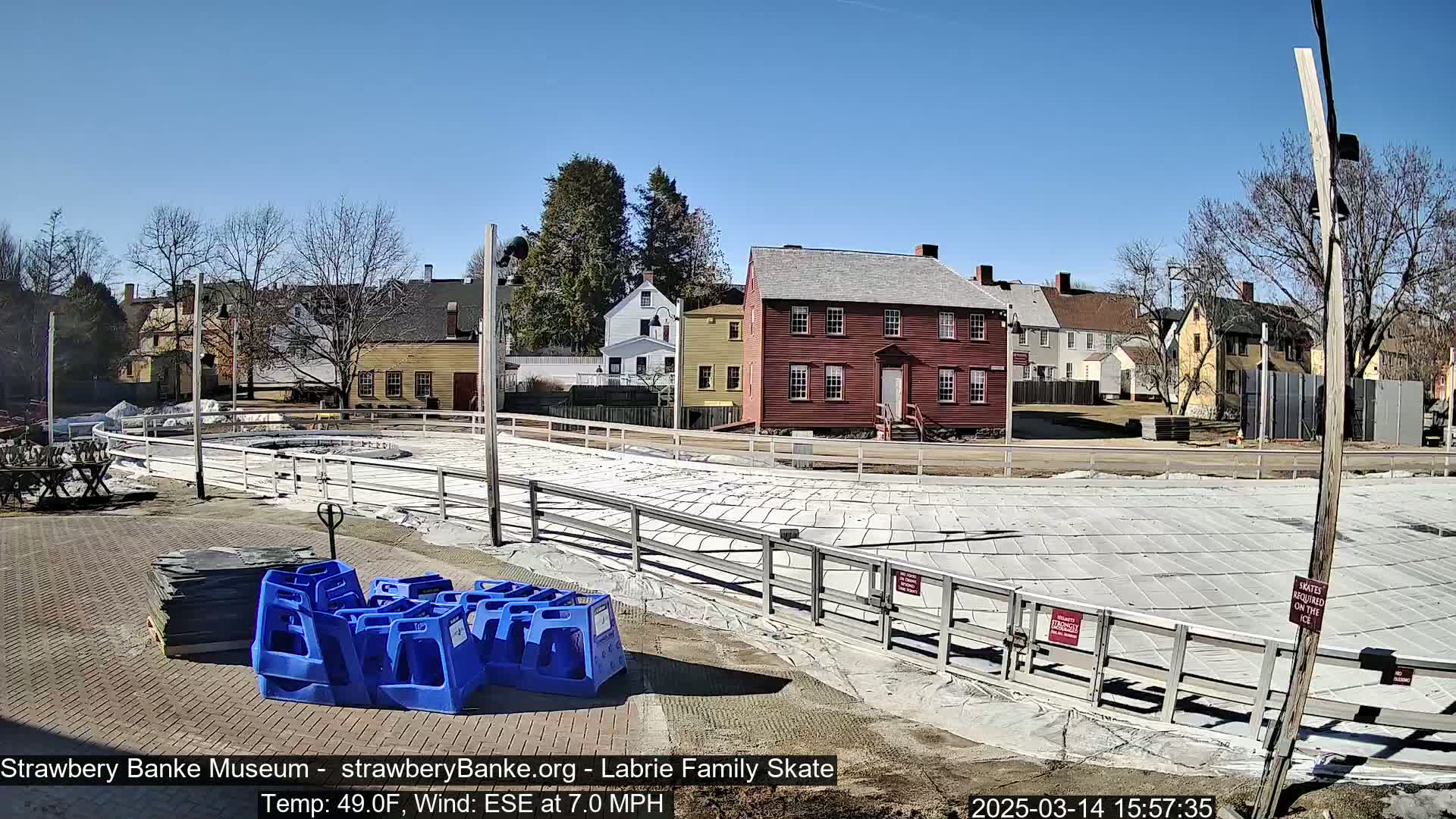 A covered outdoor skating rink surrounded by historical buildings under a clear blue sky, with a light wind.