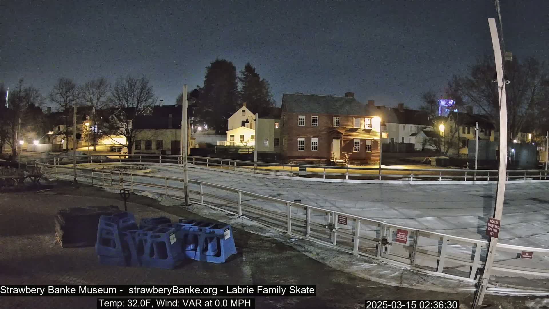 An empty outdoor ice rink is surrounded by historic-looking buildings under a dark, clear night sky.