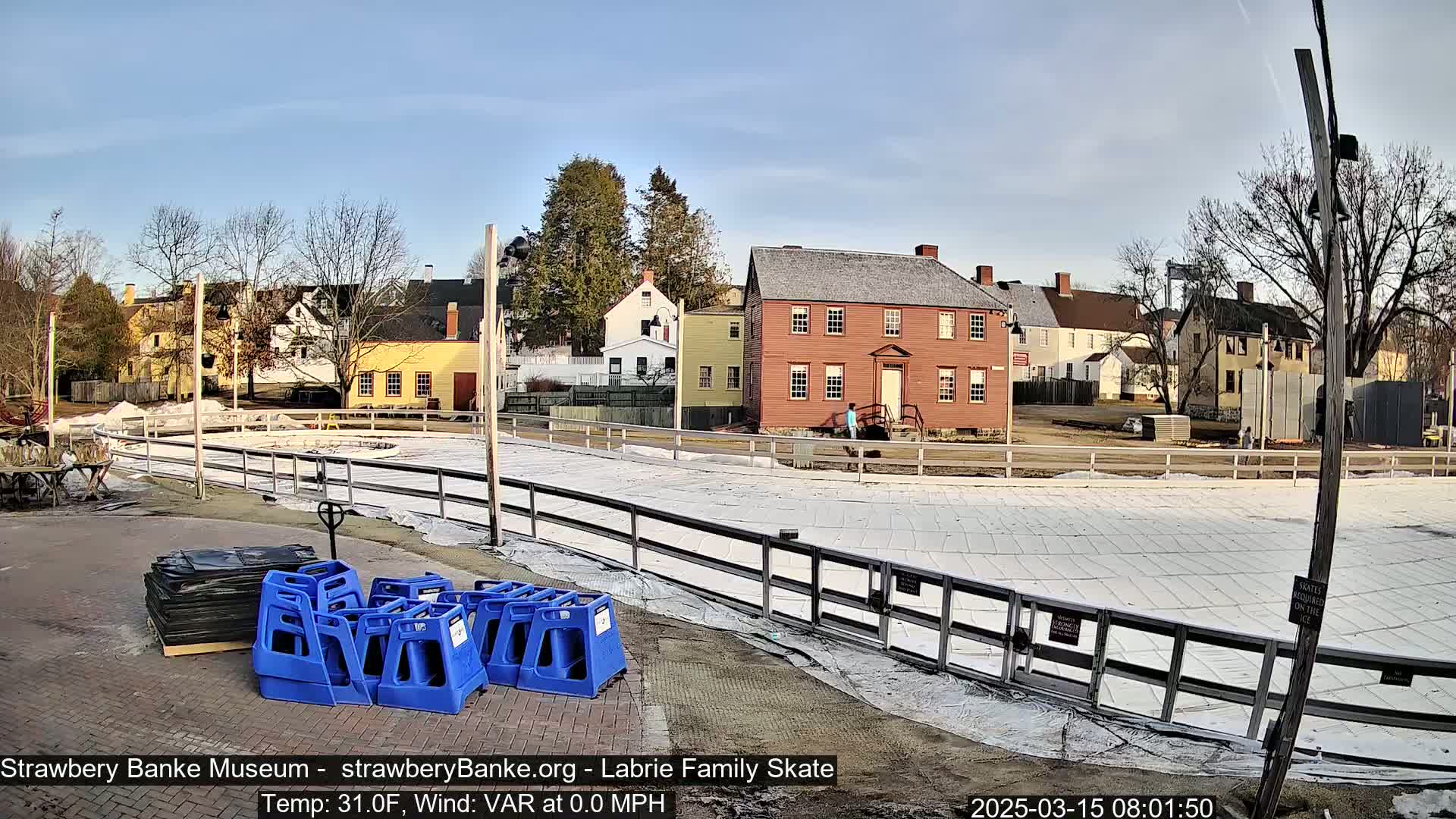 A covered outdoor ice rink sits in front of several historic-looking buildings on a sunny day with a calm wind.
