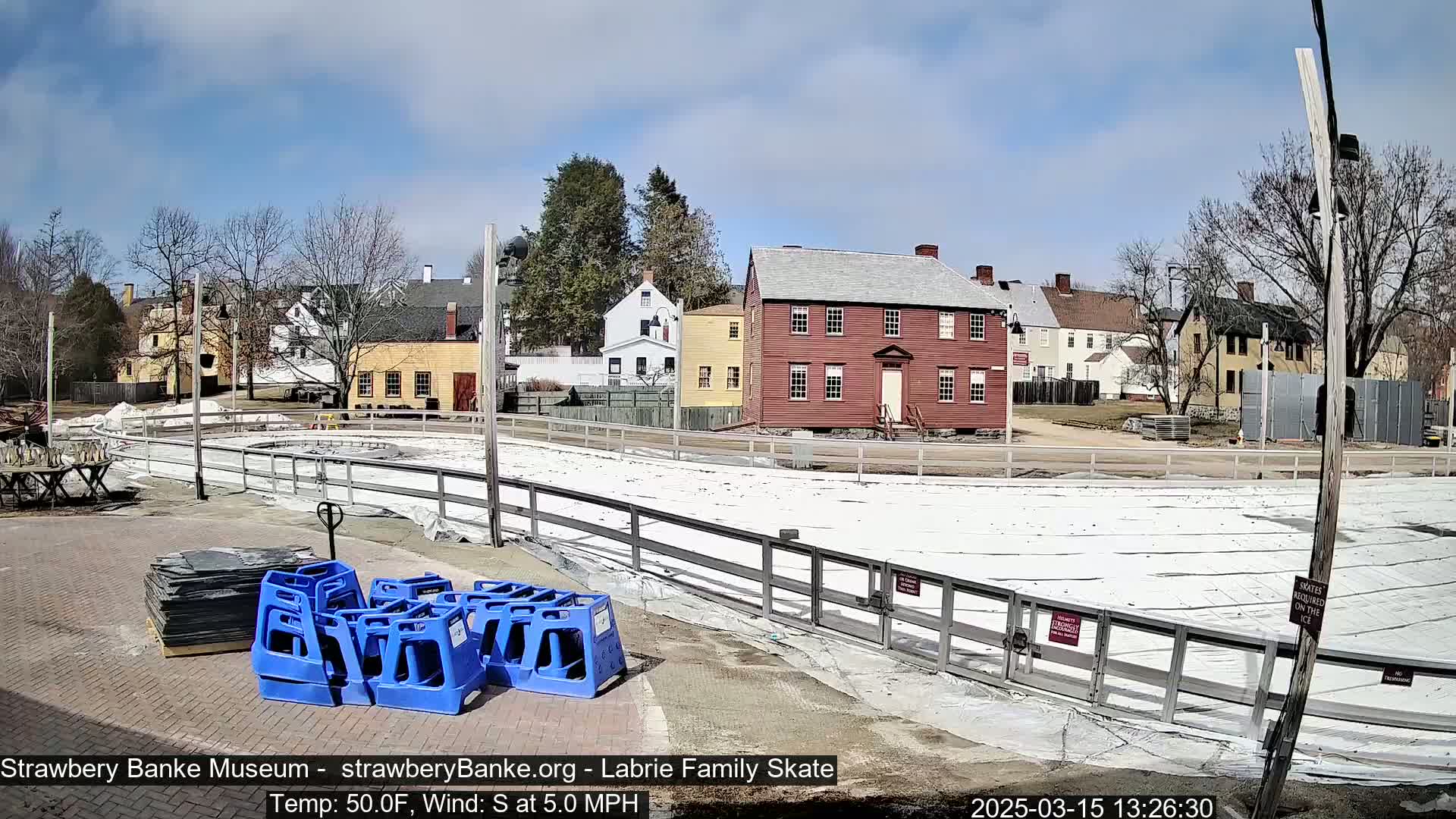 A partially covered outdoor ice rink sits in front of several historic-looking buildings on a sunny day with a light south wind.