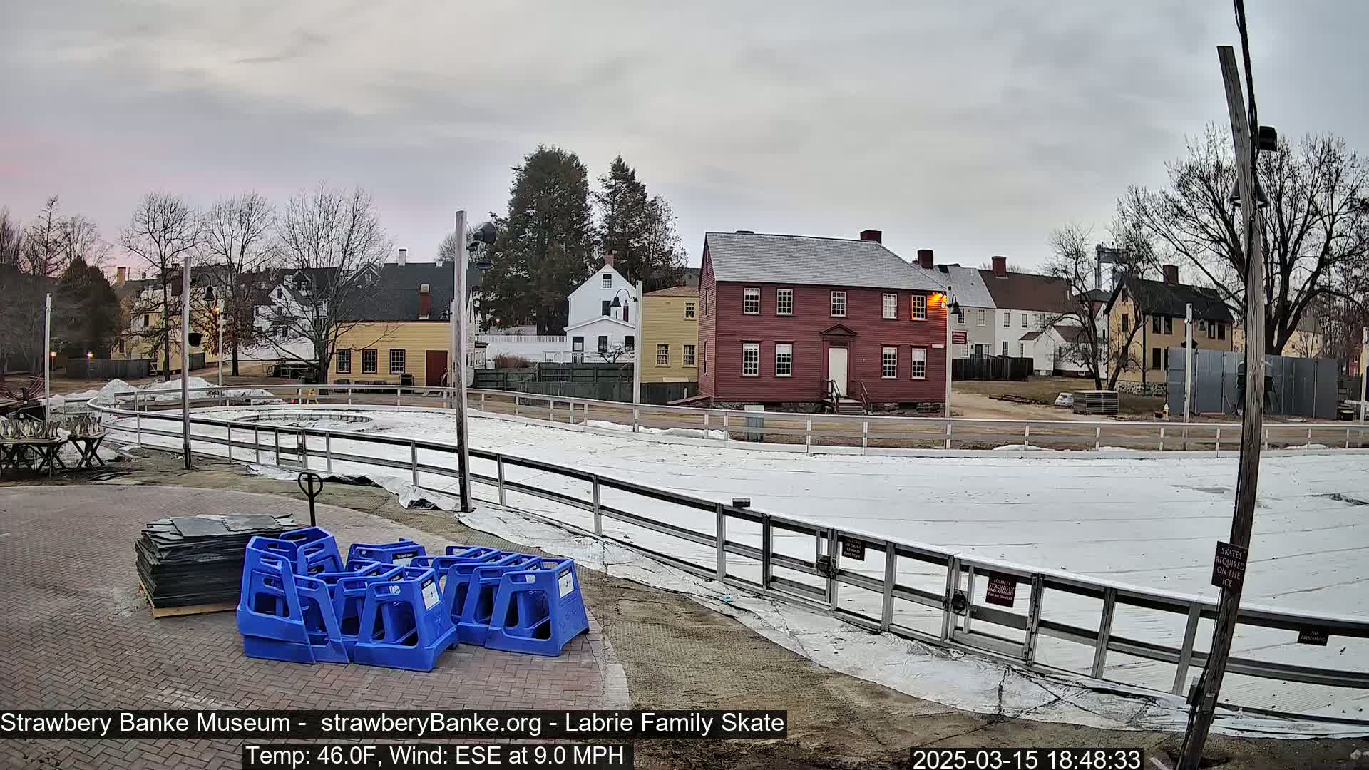 A covered outdoor ice rink sits in front of several historic buildings under an overcast sky.