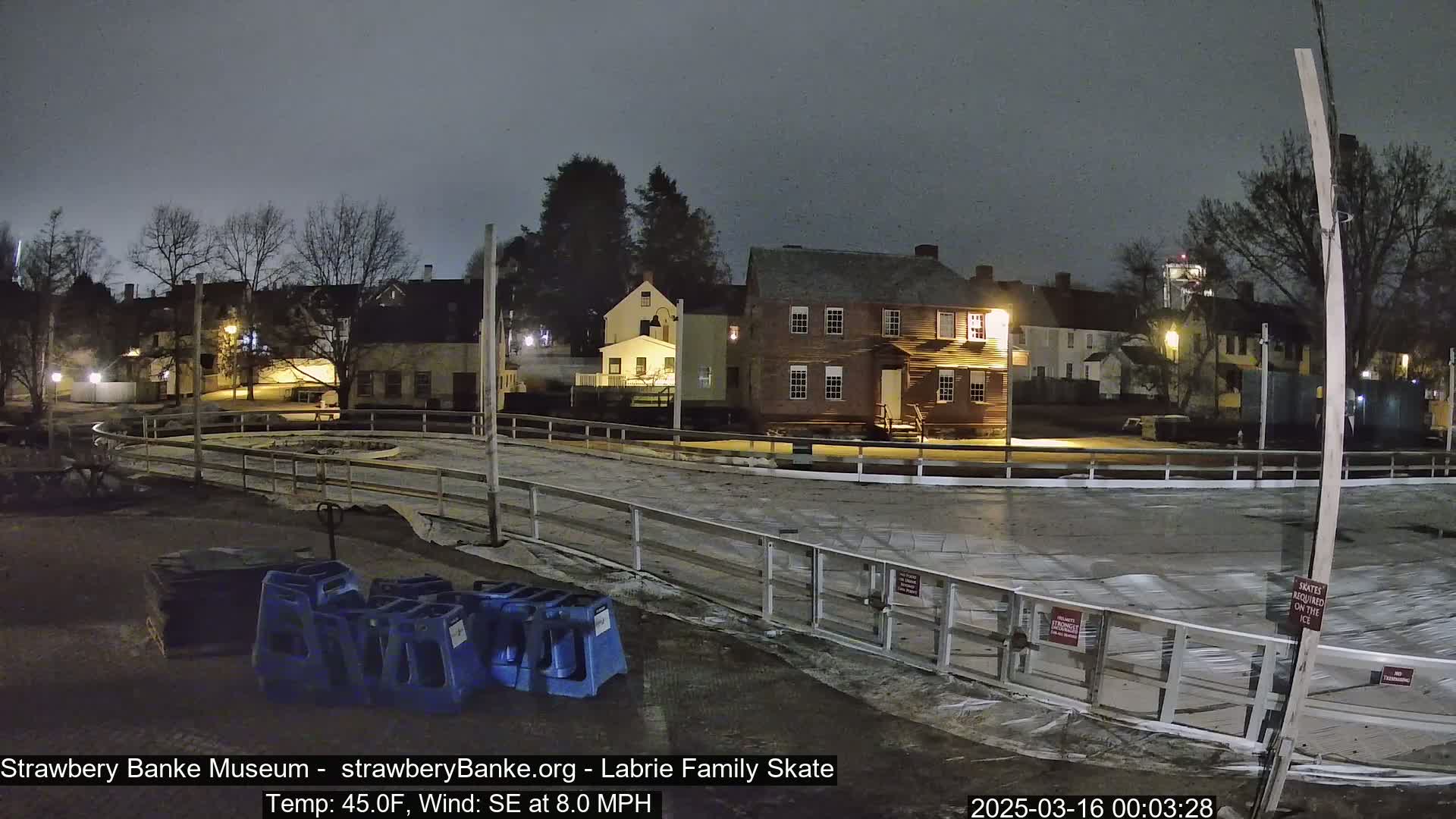 At night, under a gray sky, a partially-lit outdoor ice rink is surrounded by historic-looking buildings, with a southeast wind blowing at 8 mph and a temperature of 45°F.