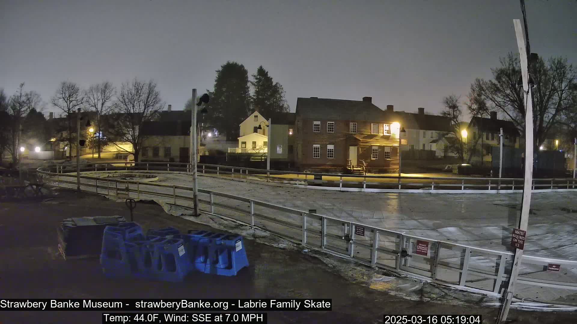 A nighttime view of an empty outdoor ice rink surrounded by historical-looking buildings under a cloudy sky.