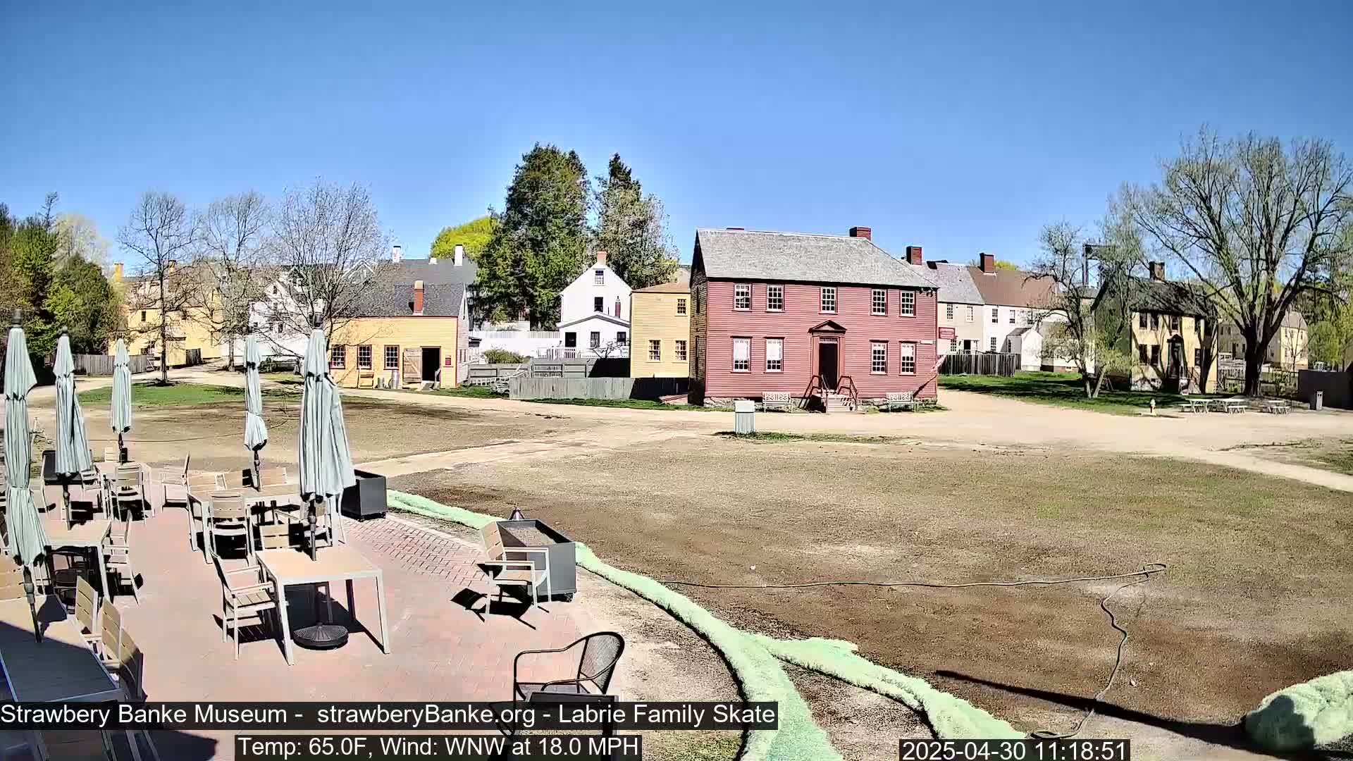 A sunny outdoor patio with tables and chairs sits before a collection of historic-looking buildings on a mostly clear day with a strong wind.
