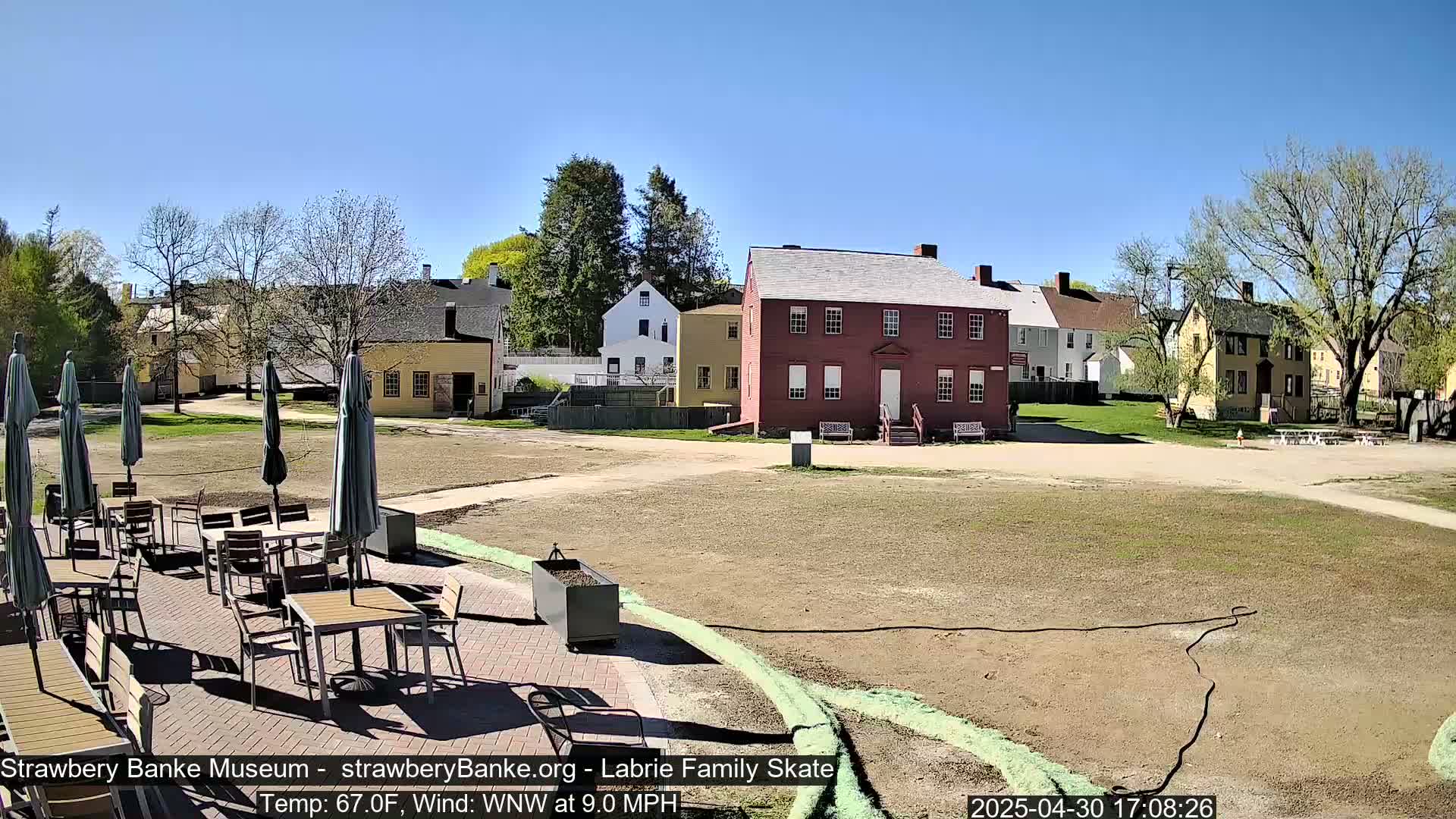 A patio with outdoor furniture sits in front of a grassy area and several historic-looking buildings on a sunny day with a light wind.