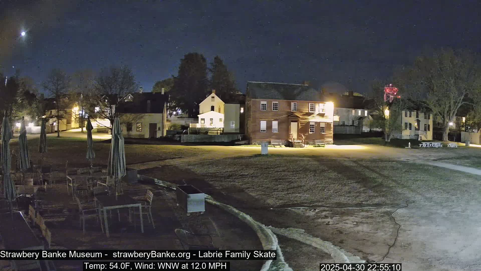 A nighttime view of a historical-looking village with several buildings, outdoor furniture, and closed umbrellas under a partly cloudy, starry sky.