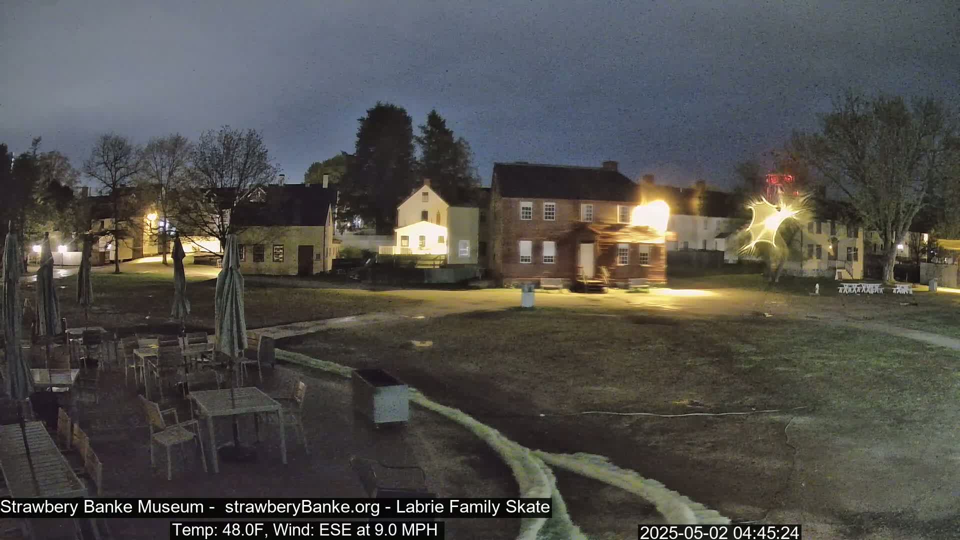 At night, under a dark sky with a 9 mph ESE wind and 48°F temperature, a courtyard scene shows several historic buildings illuminated, outdoor furniture under umbrellas, and a grassy area.