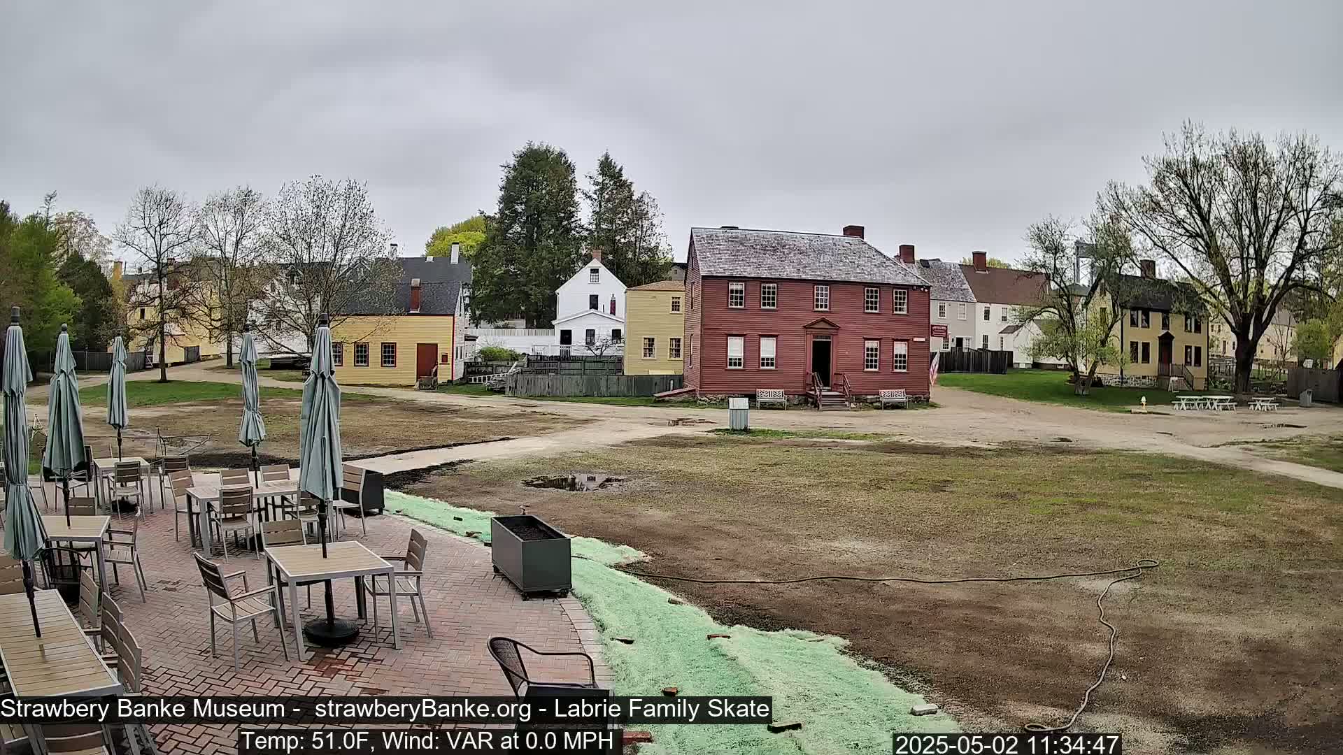A patio with tables and umbrellas is situated in front of several historic-looking buildings on an overcast day.