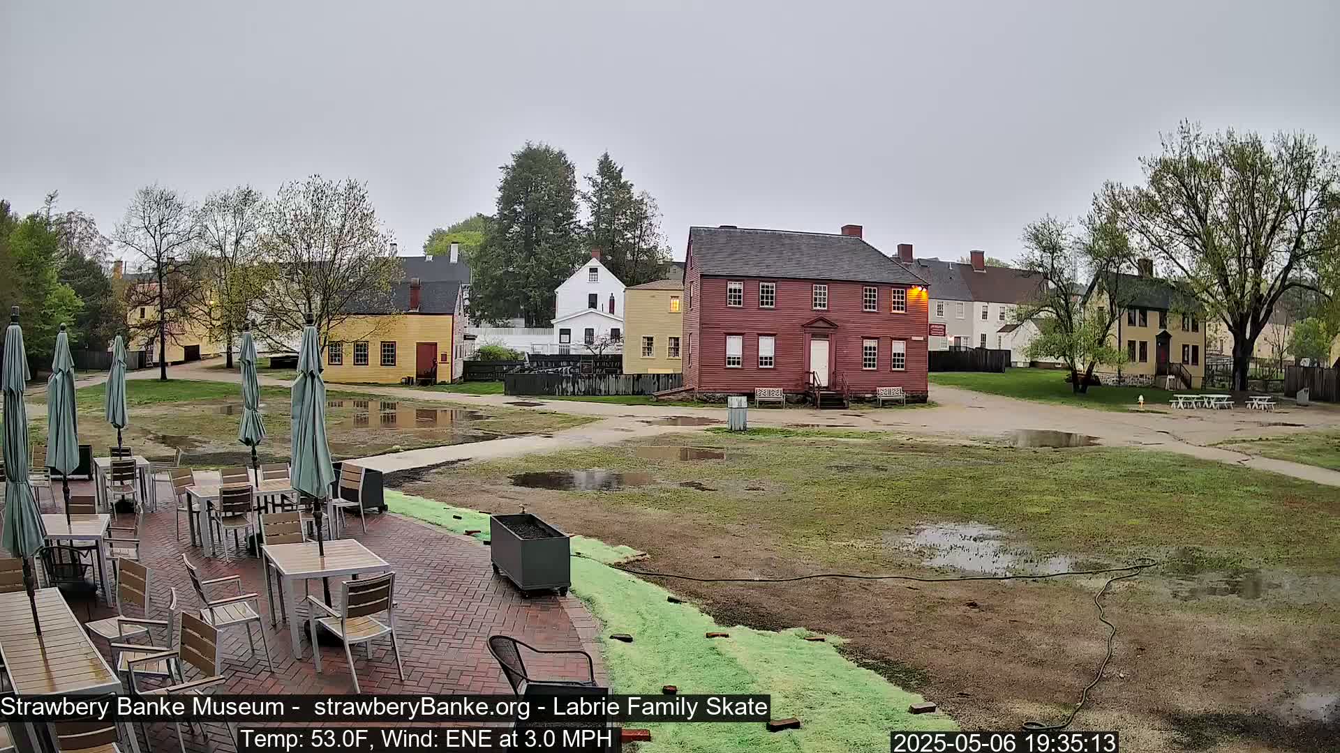 A patio with tables and umbrellas sits beside a wet grassy area and several historic-looking buildings under an overcast sky.