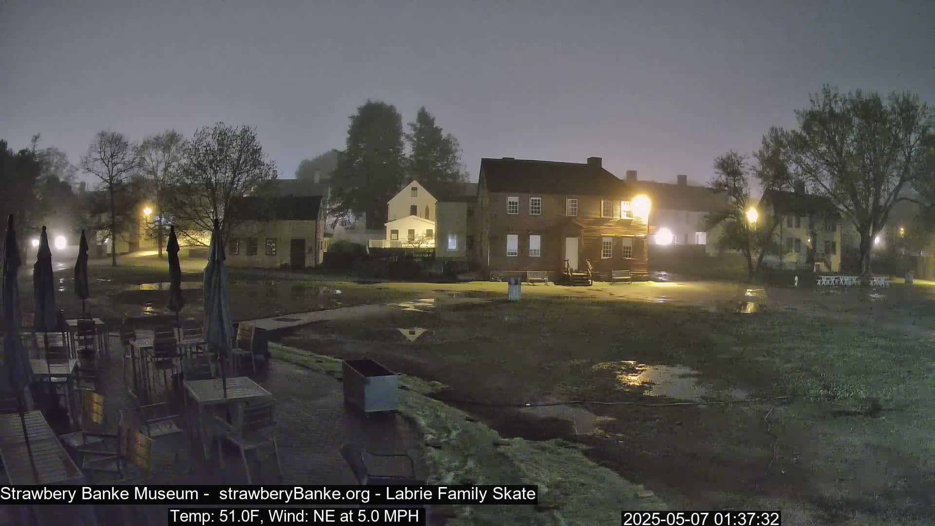 A foggy, nighttime scene shows several colonial-style buildings and a wet, open area with outdoor furniture and umbrellas.