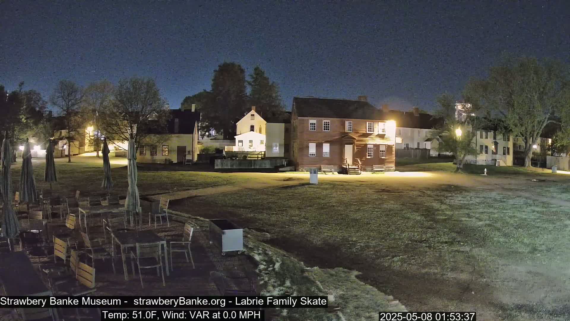Under a dark, clear night sky, several colonial-style buildings are illuminated, surrounding a grassy area with outdoor furniture and closed umbrellas.