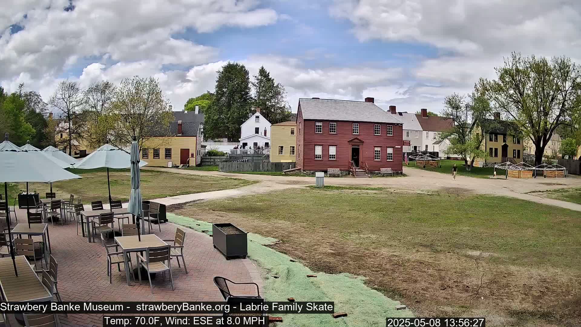 A patio with tables and umbrellas overlooks a grassy area and several historic-looking buildings under a partly cloudy sky.