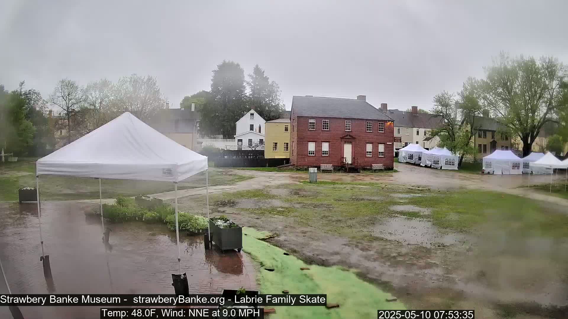 A rain-soaked courtyard with several white event tents and historic buildings, under a gray sky.