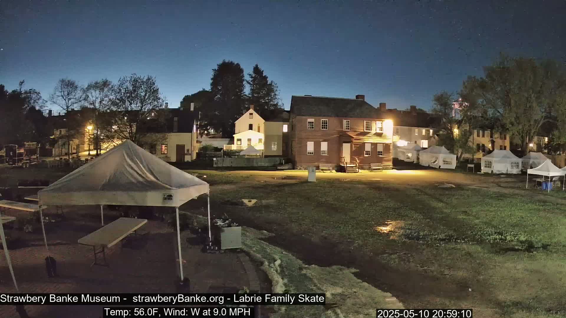Under a clear, dark night sky, several tents and historic-looking buildings are illuminated in an outdoor area, which appears damp and possibly muddy.