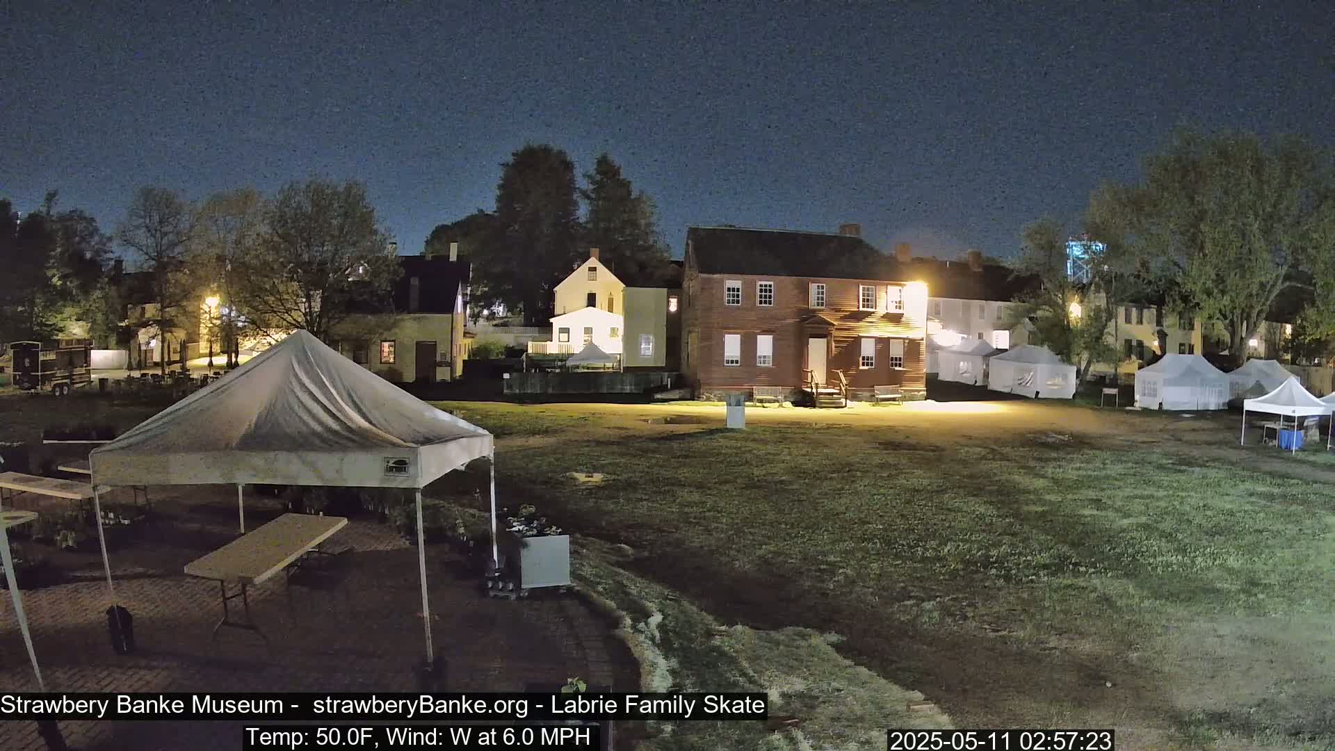At night, several tents are set up on a grassy field in front of a row of historic-looking buildings under a clear, dark sky.