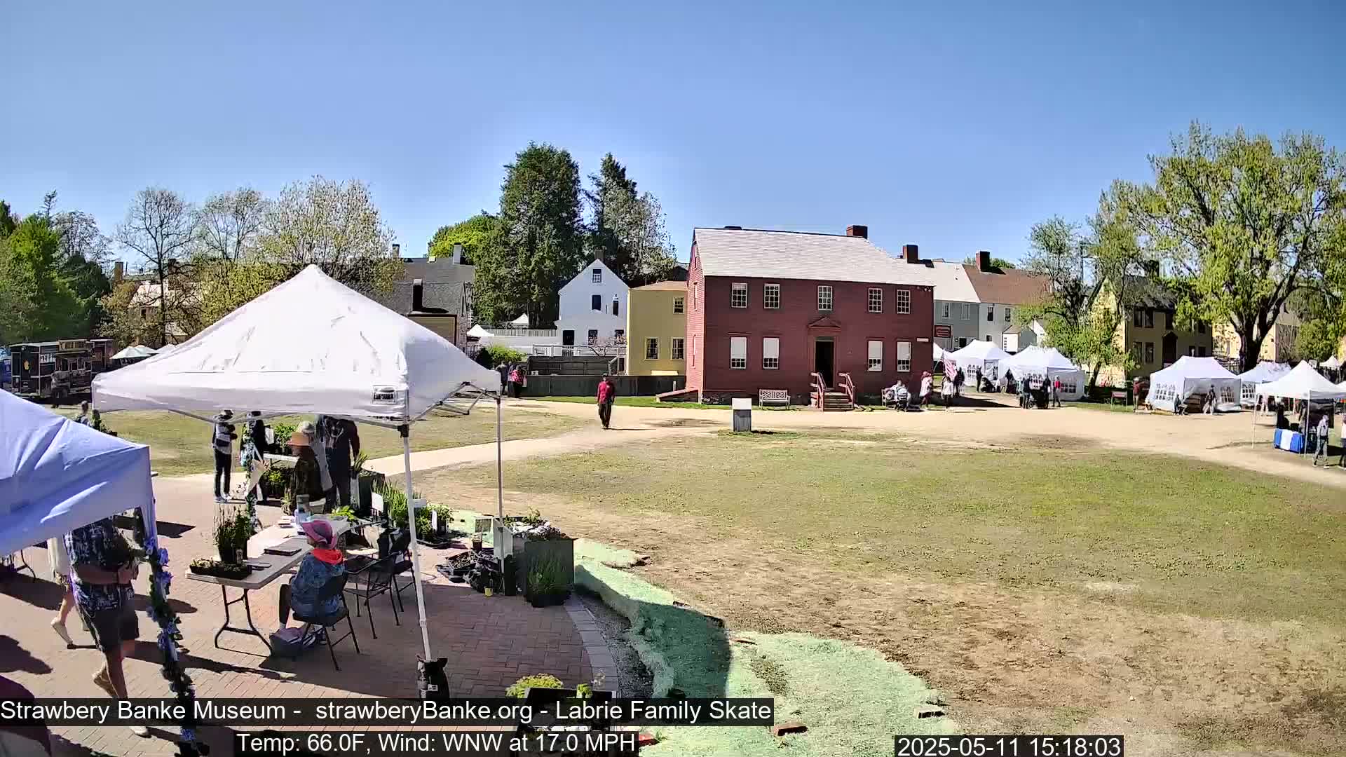 A sunny outdoor scene shows several white tents set up on a grassy area in front of historical-looking buildings, with people milling about.