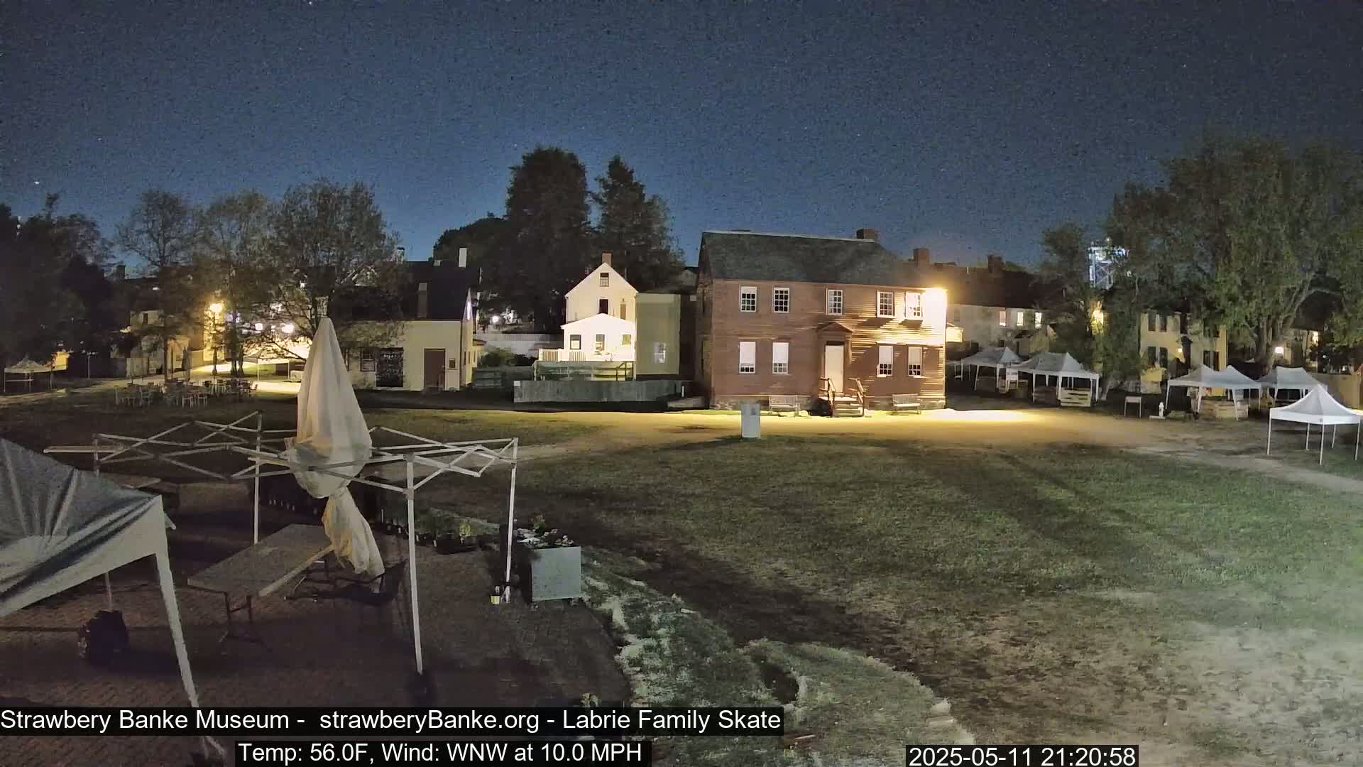 A nighttime view of a grassy area with several buildings, some tents, and outdoor furniture under a clear, dark sky.