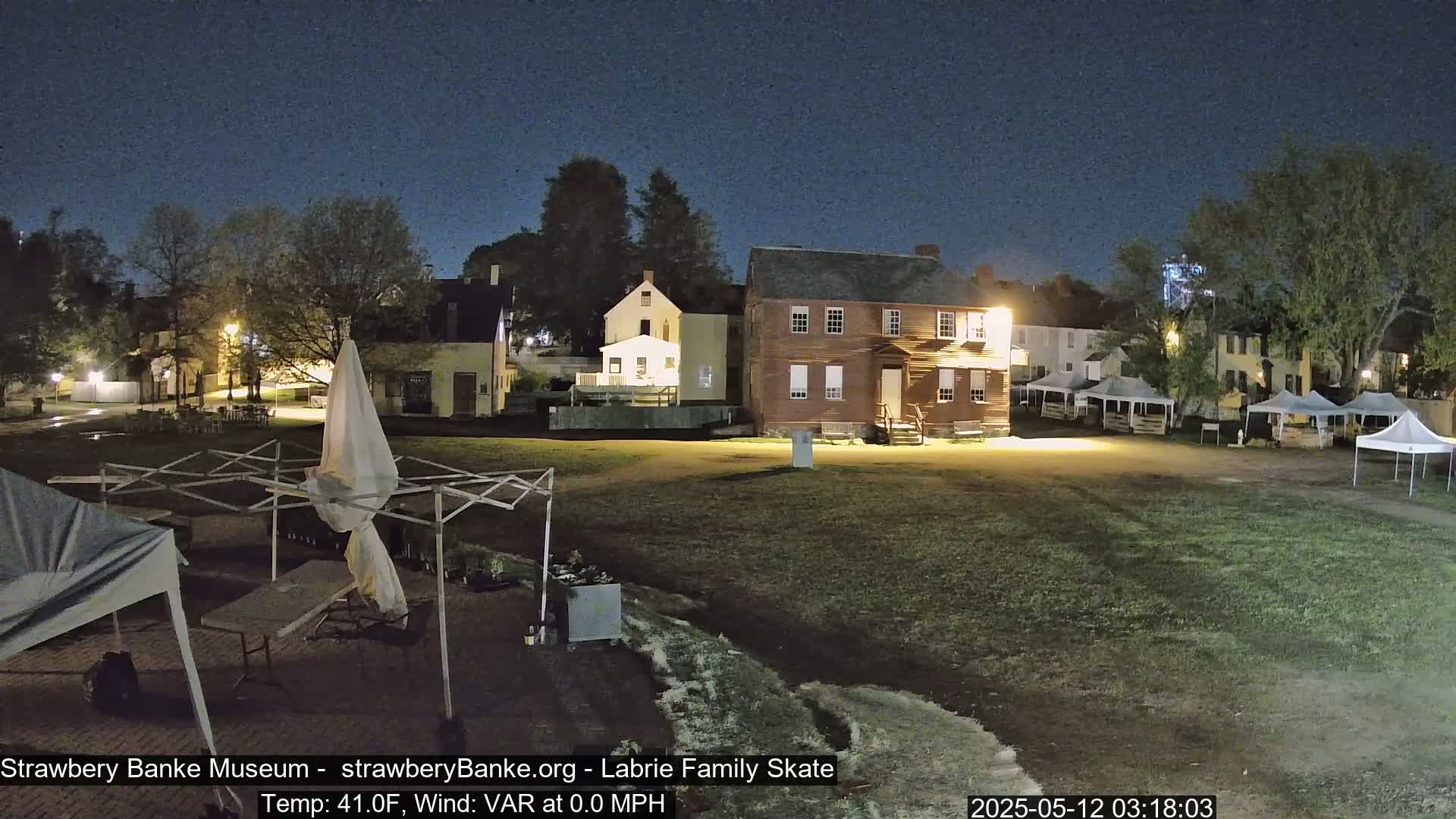 At night, several historical buildings are illuminated, surrounded by a grassy area with numerous canopies and tents.
