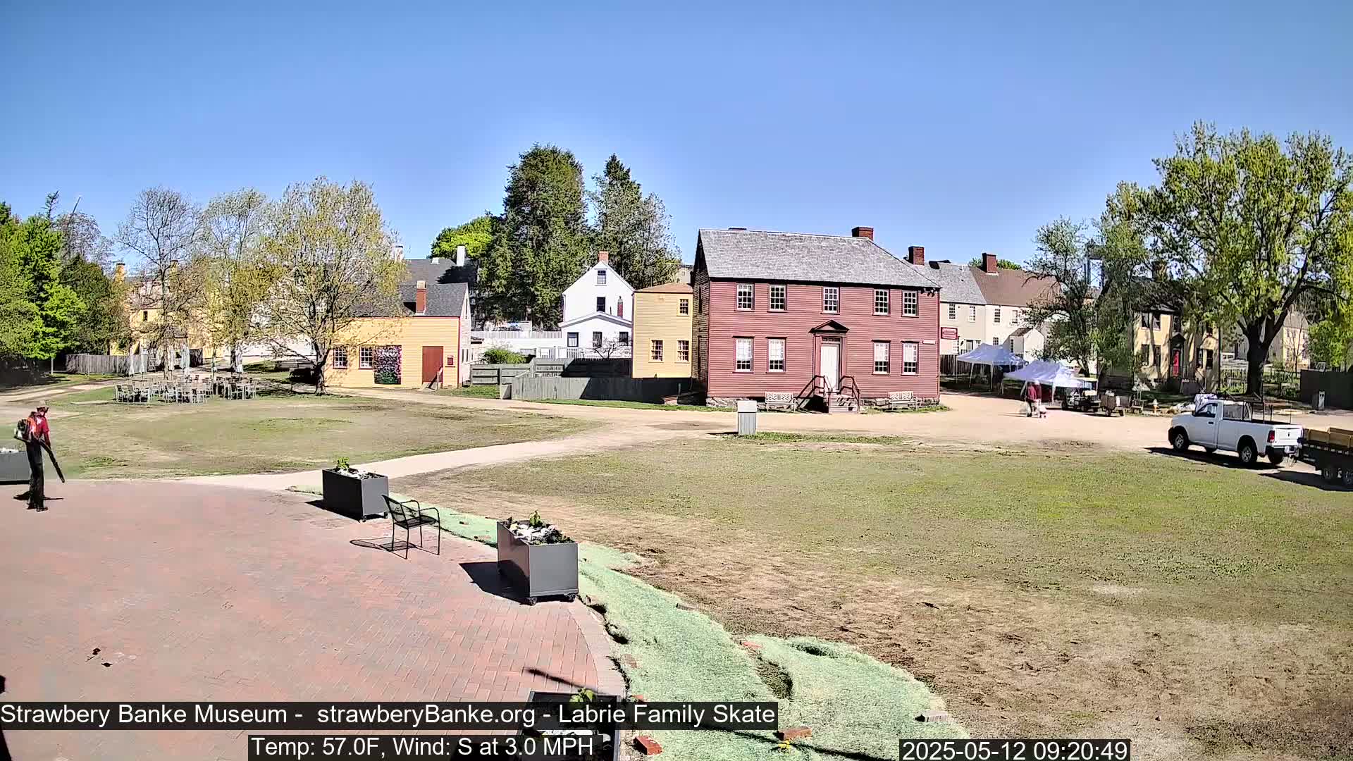 A sunny outdoor scene shows several colonial-style buildings, a person using a leaf blower, and a pickup truck parked in a grassy area.