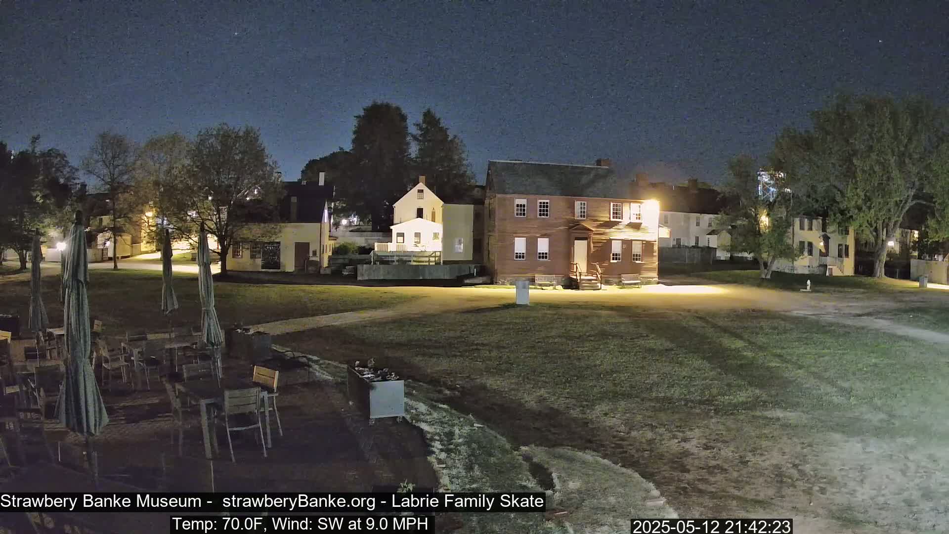 A nighttime scene shows several historical-looking buildings illuminated under a clear, dark sky, with an outdoor seating area containing several tables and chairs in a grassy area in front of them.