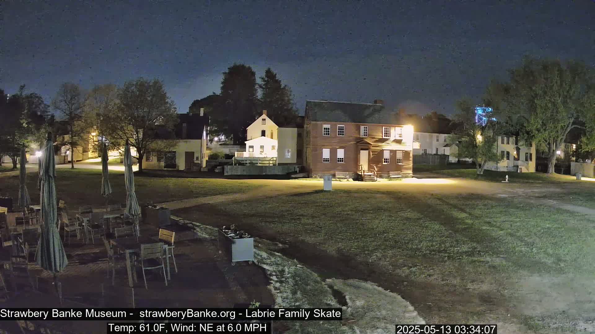 A nighttime scene shows several historic-looking buildings surrounded by a grassy area with outdoor seating under umbrellas, a slight northeast wind, and a temperature of 61 degrees Fahrenheit.