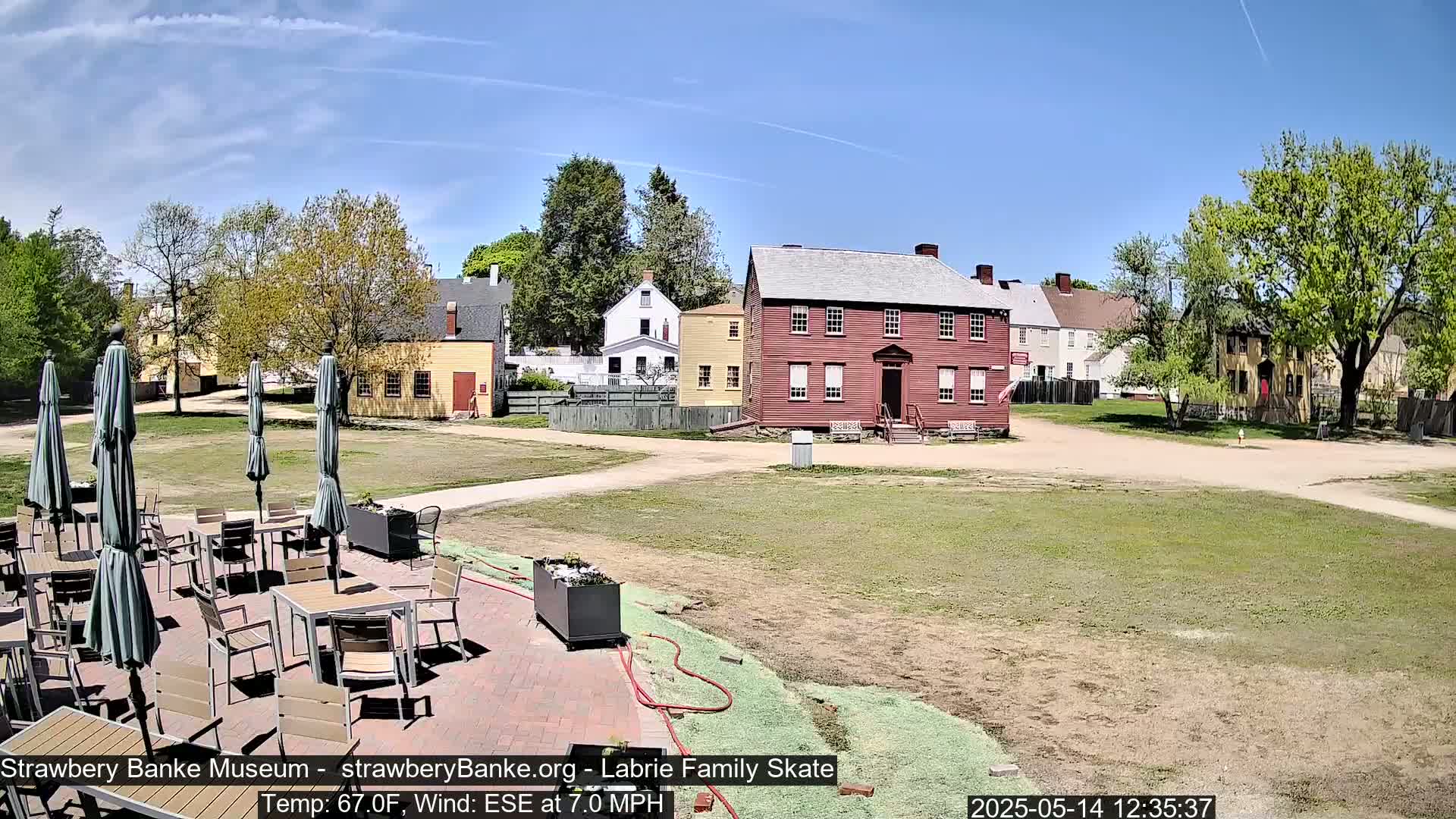 A sunny outdoor scene shows a patio with tables and umbrellas in front of several historic-looking buildings on a grassy area with a slight wind.