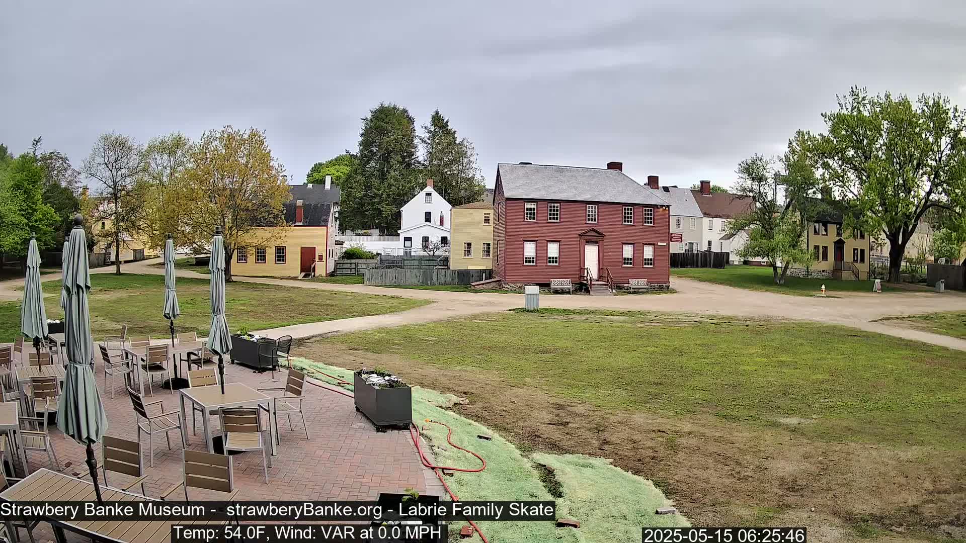 A patio with tables and umbrellas sits beside a grassy area, overlooking several historic-looking buildings under an overcast sky.