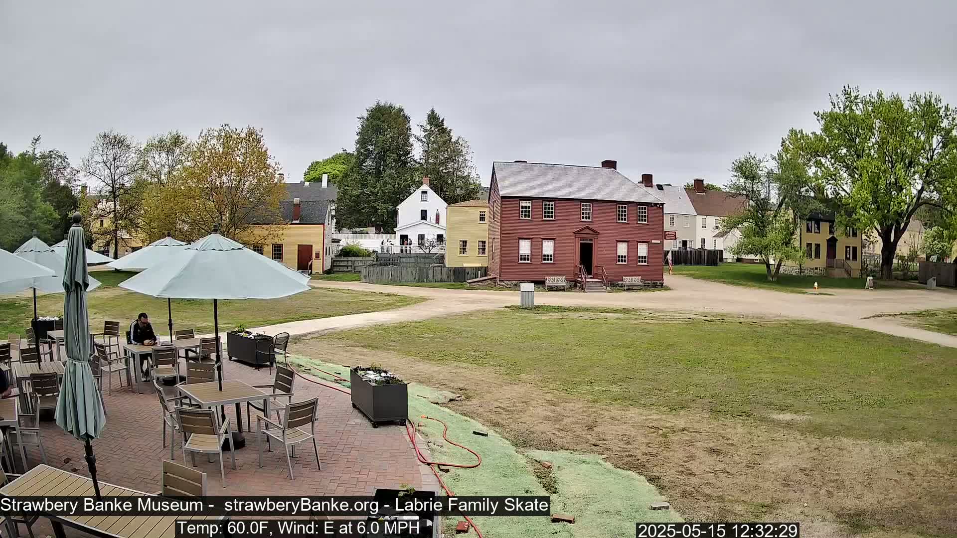 A person sits at an outdoor cafe table under a patio umbrella, overlooking a grassy area with several historic-looking buildings in the background on an overcast day.