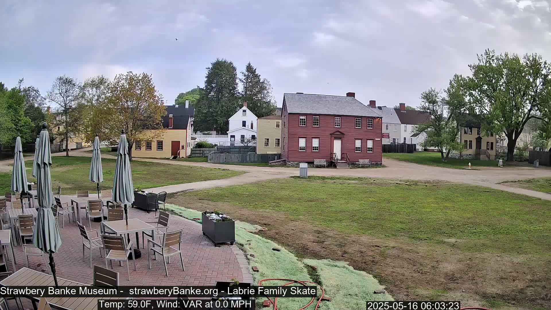 An outdoor patio with tables and umbrellas overlooks a grassy area with several historic-looking buildings under a partly cloudy sky.