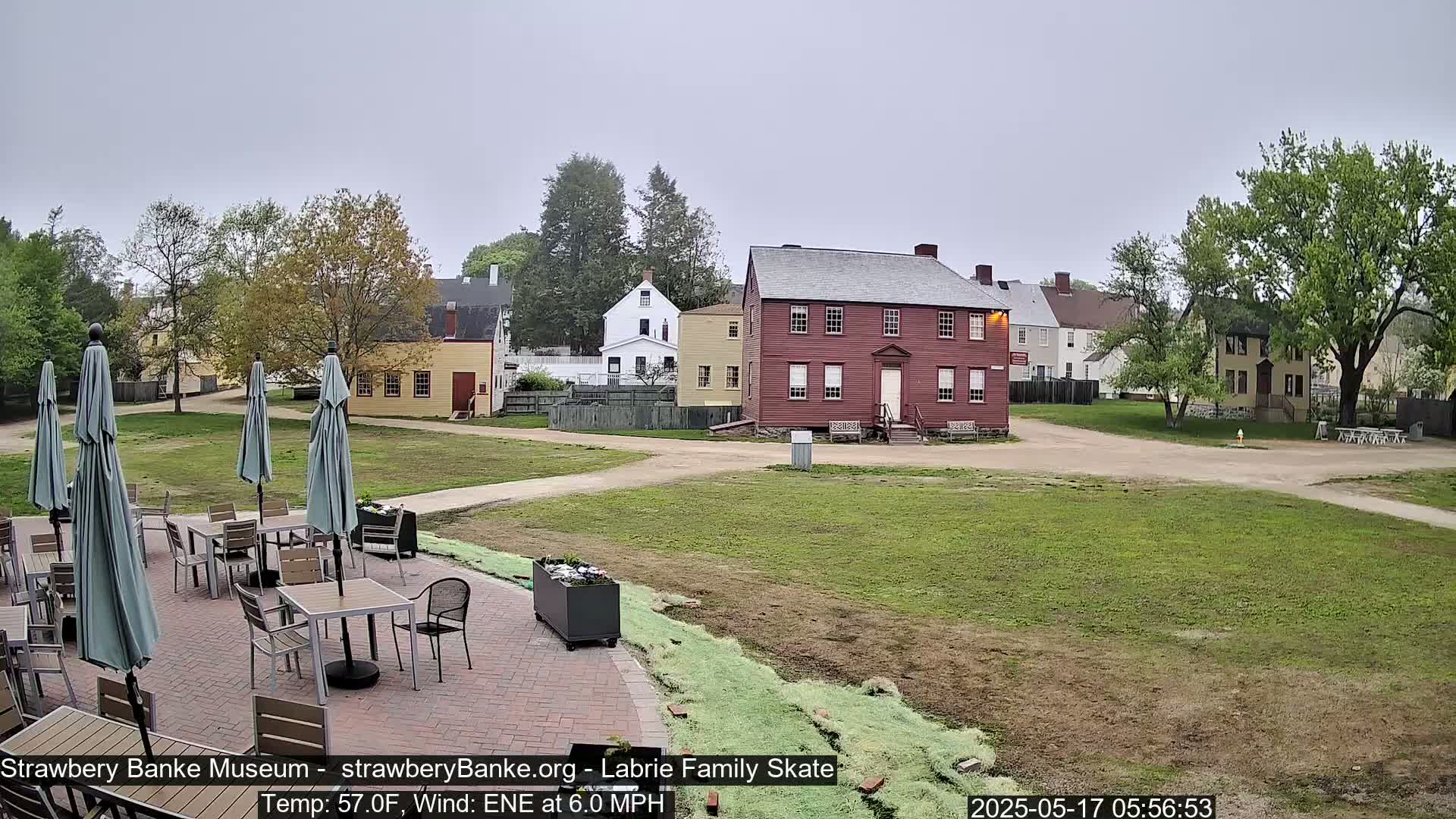 An outdoor patio with tables and umbrellas sits before several historic buildings on a grassy area under an overcast sky.