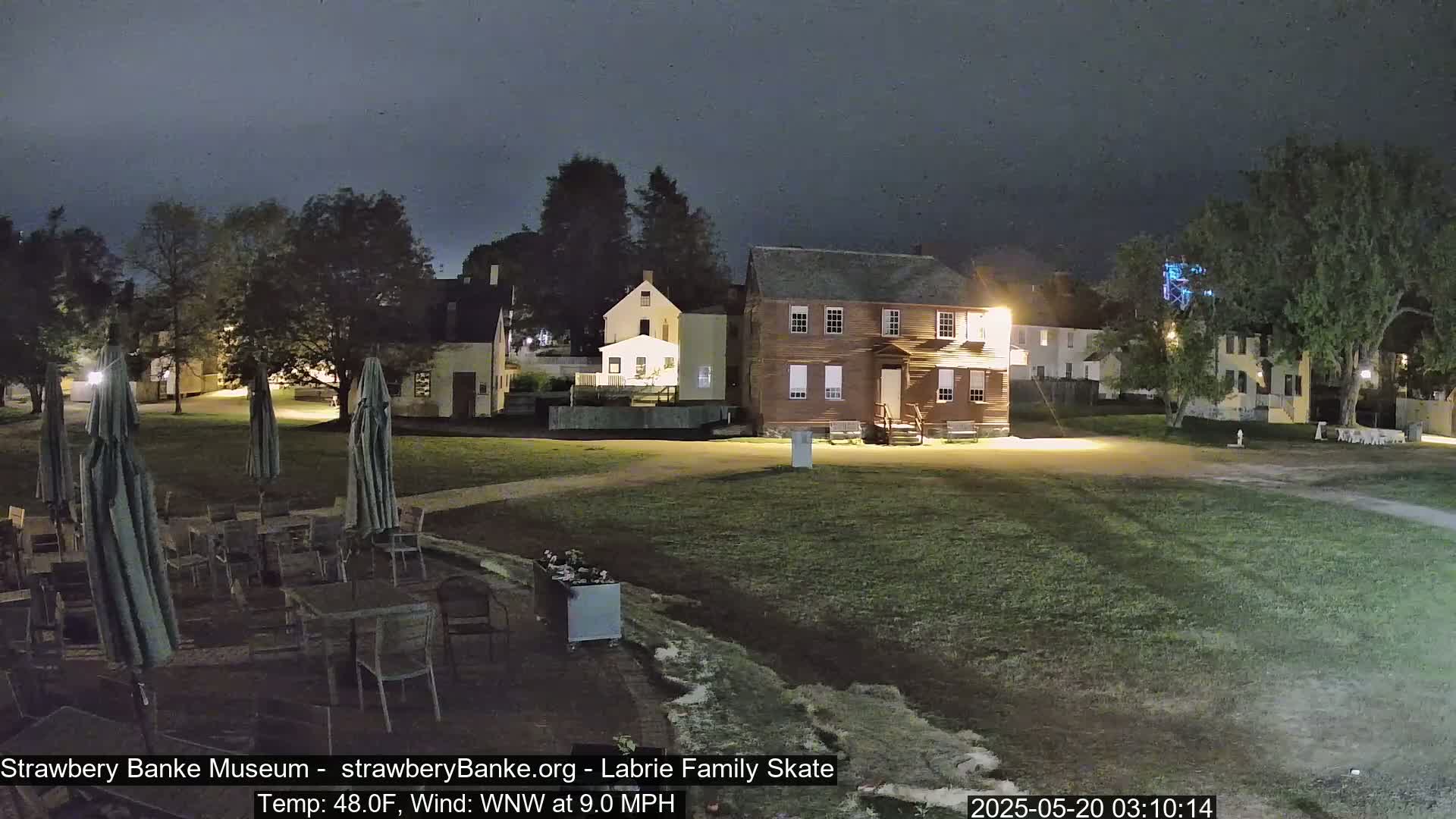 A nighttime view of a grassy area with several historical buildings and outdoor patio furniture under a dark sky.